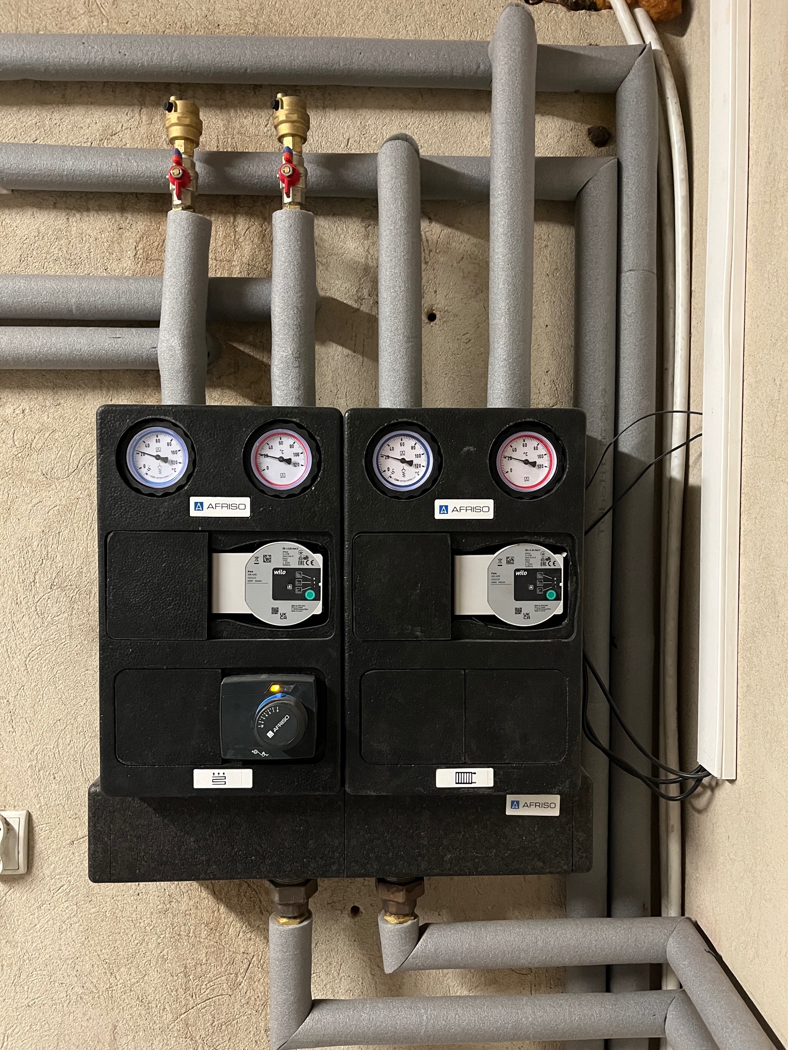 Dwa czarne moduły pompy ciepła AFRISO z manometrami i pompami obiegowymi Wilo, zamontowane na ścianie z rurami izolowanymi szarą pianką.