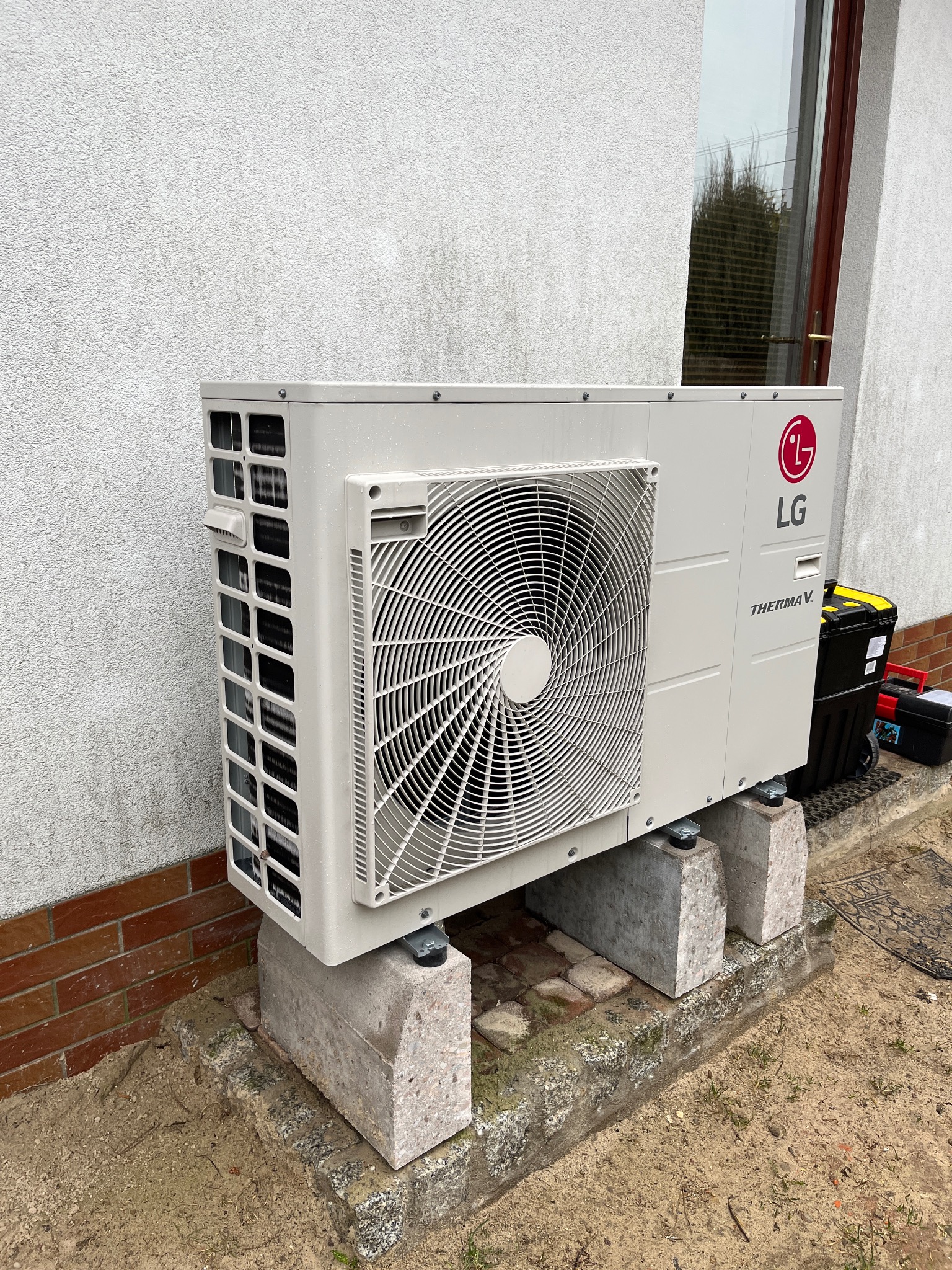 Zewnętrzna jednostka pompy ciepła LG Therma V, zamontowana na betonowych wspornikach przy ścianie budynku, obok skrzynka z narzędziami.