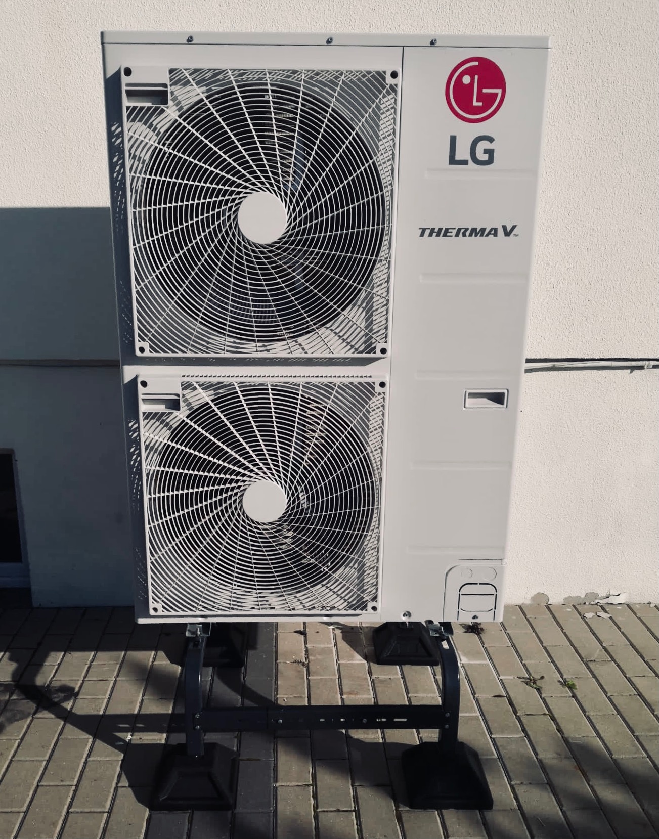 Zewnętrzna jednostka pompy ciepła LG Therma V, zamontowana na czarnej metalowej podstawie, na tle jasnej, teksturowanej ściany i chodnika z szarej kostki brukowej.