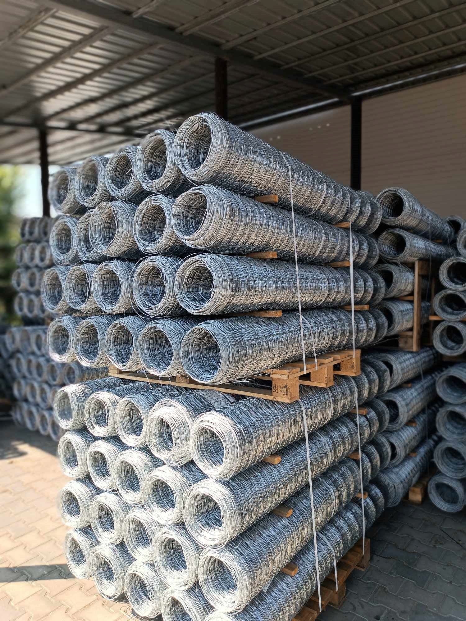 Stos rolek siatki ogrodzeniowej ocynkowanej, ułożonych na drewnianych paletach pod zadaszeniem magazynu.