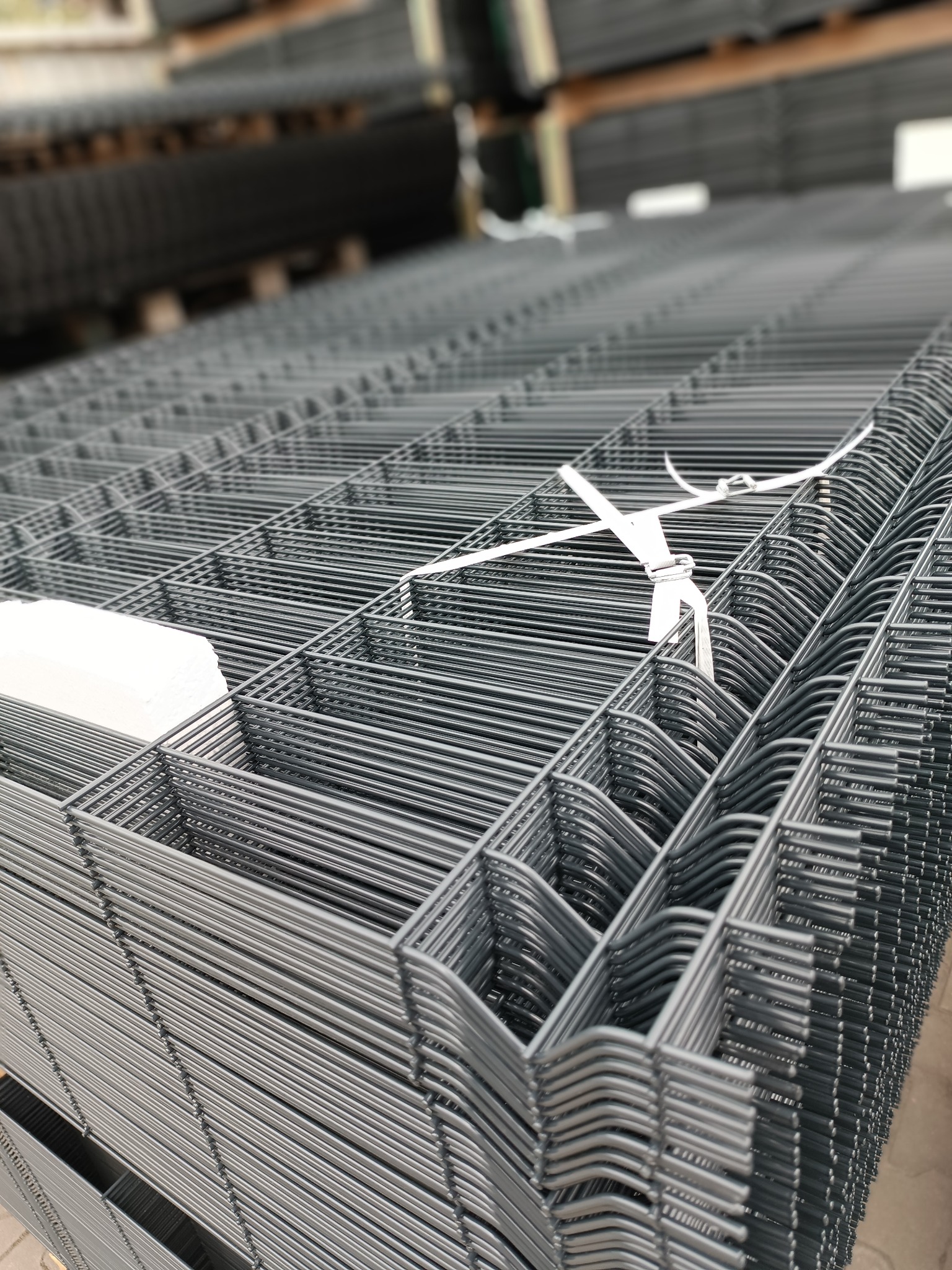 Stos szarych paneli ogrodzeniowych zgrzewanych, zabezpieczonych białą taśmą, ułożonych na drewnianych paletach, widok z góry.