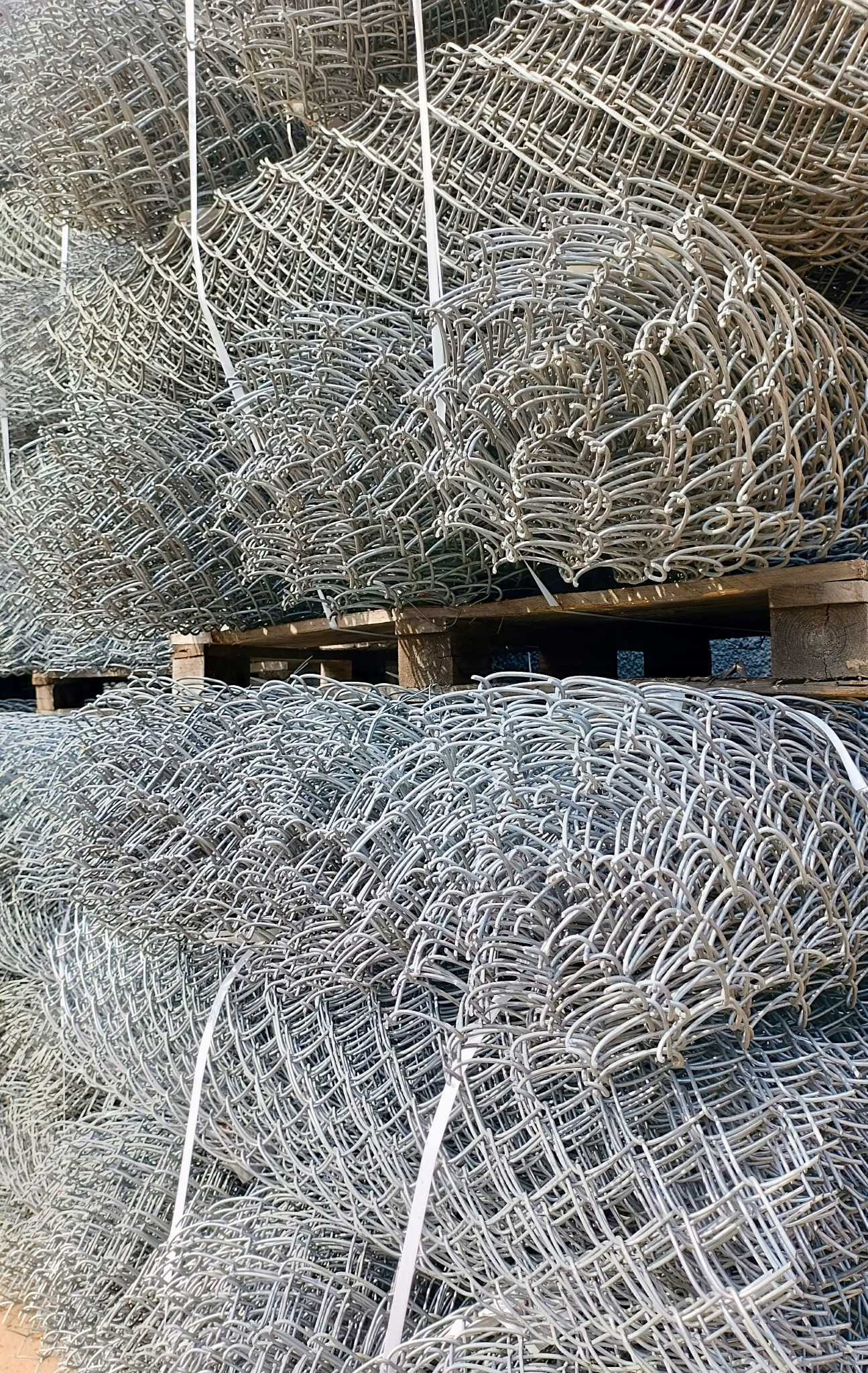 Stosy zrolowanej, ocynkowanej siatki ogrodzeniowej na drewnianych paletach, zabezpieczone białymi taśmami.