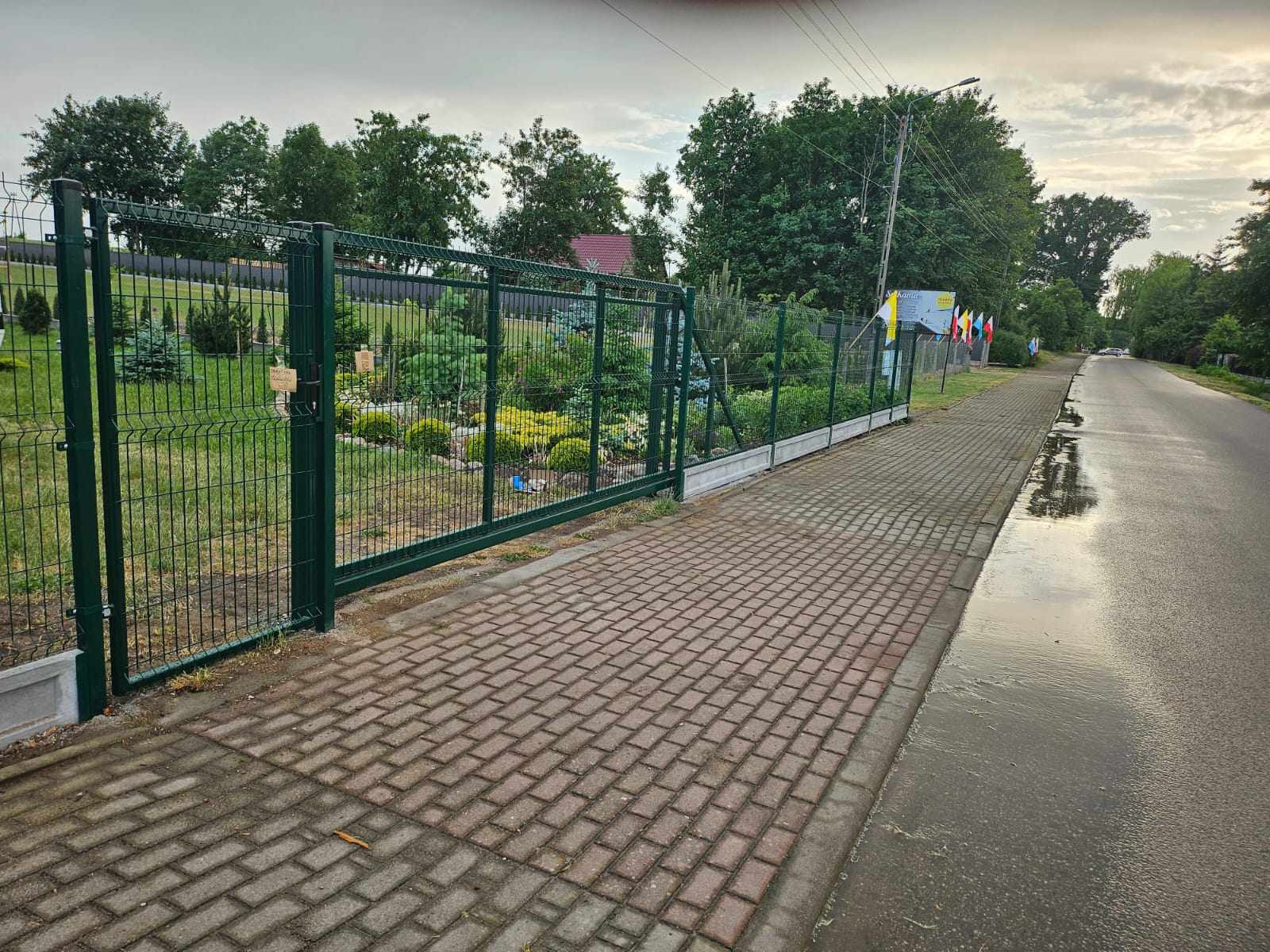 Otwarta, zielona brama z paneli ogrodzeniowych z widokiem na zadbany ogród, betonowy murek i chodnik z kostki brukowej wzdłuż mokrej jezdni.