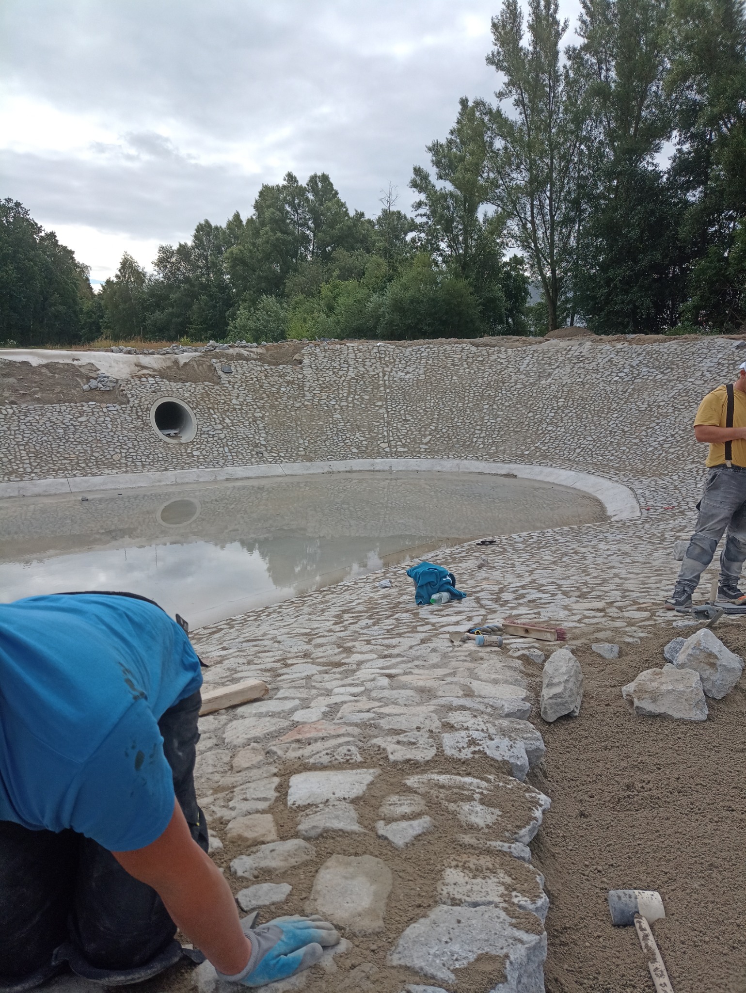 Budowa oczka wodnego z kamienną obudową w trakcie realizacji, widoczni pracownicy i narzędzia, w tle zieleń drzew.