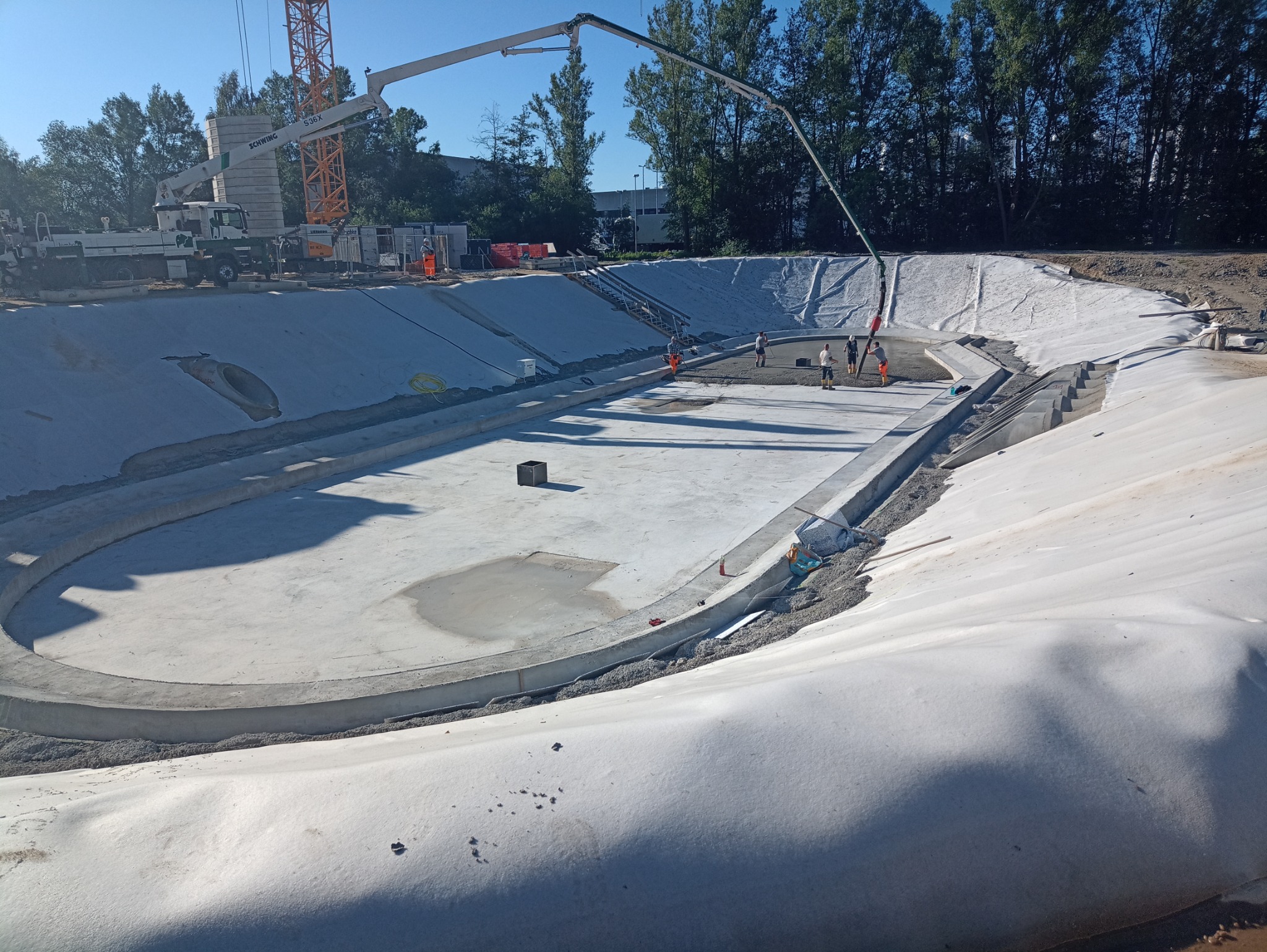 Budowa zbiornika retencyjnego: wylana betonowa podstawa i ściany, wyłożone geowłókniną, z użyciem pompy do betonu i obecnością ekipy budowlanej.