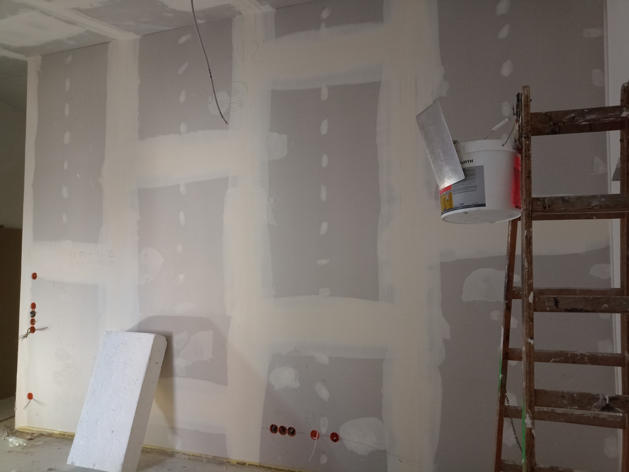 Ściana z płyt gipsowo-kartonowych w trakcie szpachlowania, widoczne miejsca łączeń i wkrętów, drabina drewniana z wiadrem farby i szpachelką, wystające kable elektryczne.