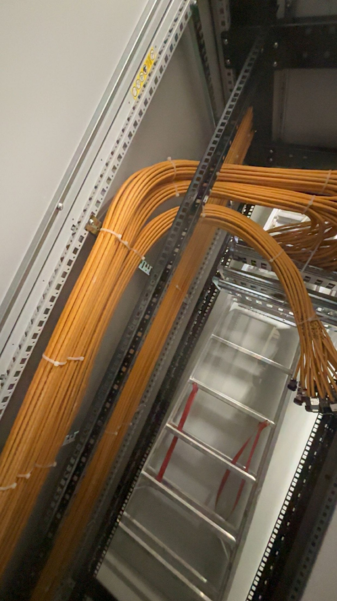 Pomarańczowe kable uporządkowane w szafie rack, zabezpieczone opaskami zaciskowymi, widoczne elementy konstrukcyjne szafy i drabinka kablowa.