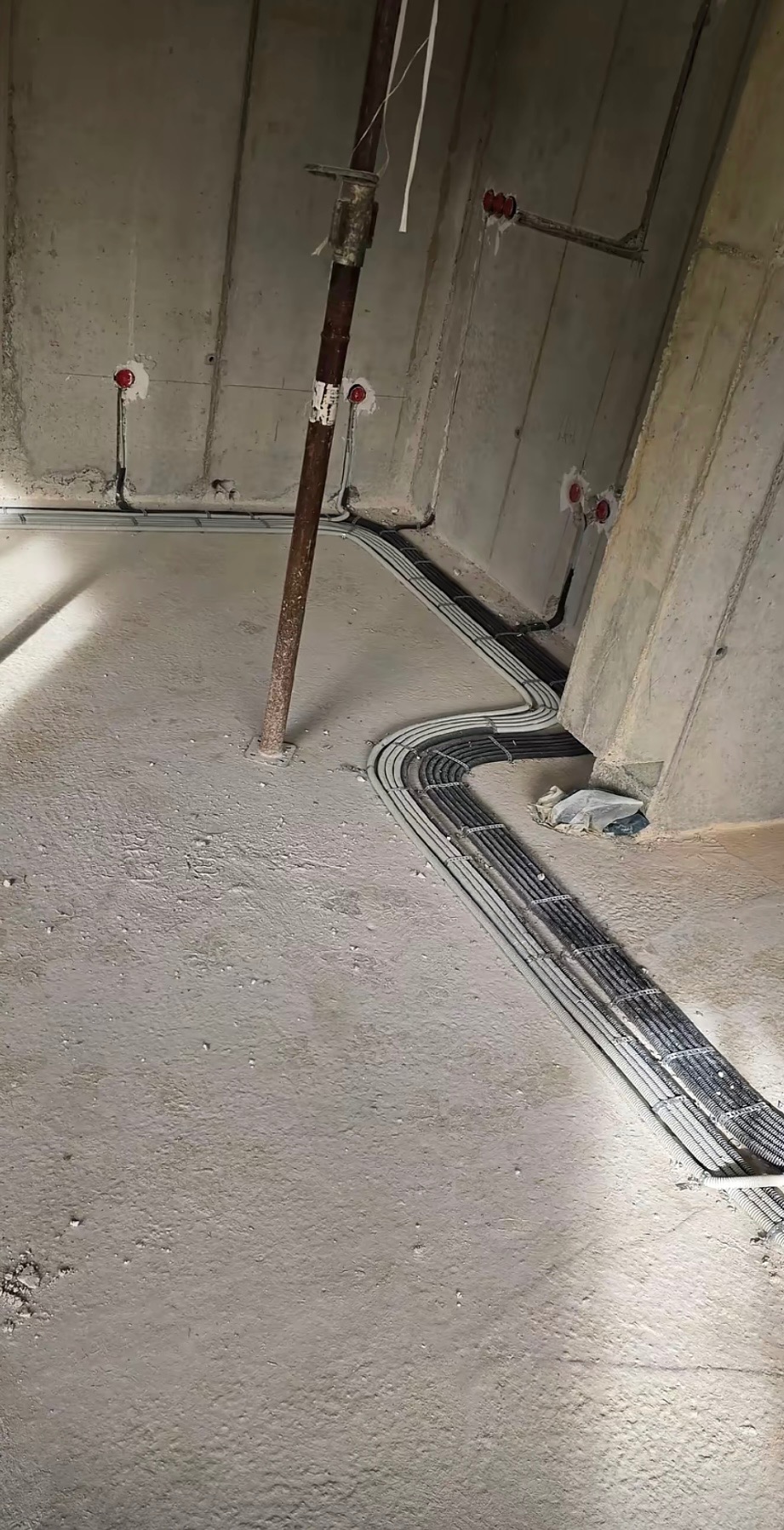 Surowy betonowy pokój w trakcie budowy z ułożonymi, szarymi rurami kablowymi wzdłuż ścian i wystającymi puszkami instalacyjnymi w ścianach.