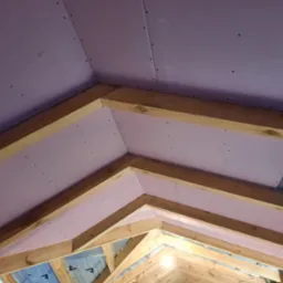 Montaż różowych płyt na drewnianej konstrukcji poddasza, widoczne wkręty i izolacja z folii aluminiowej.