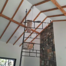 Wnętrze w trakcie prac wykończeniowych: widoczne drewniane belki stropowe, fragment ściany wyłożonej kamieniem dekoracyjnym oraz rusztowanie z drewnianymi podestami.