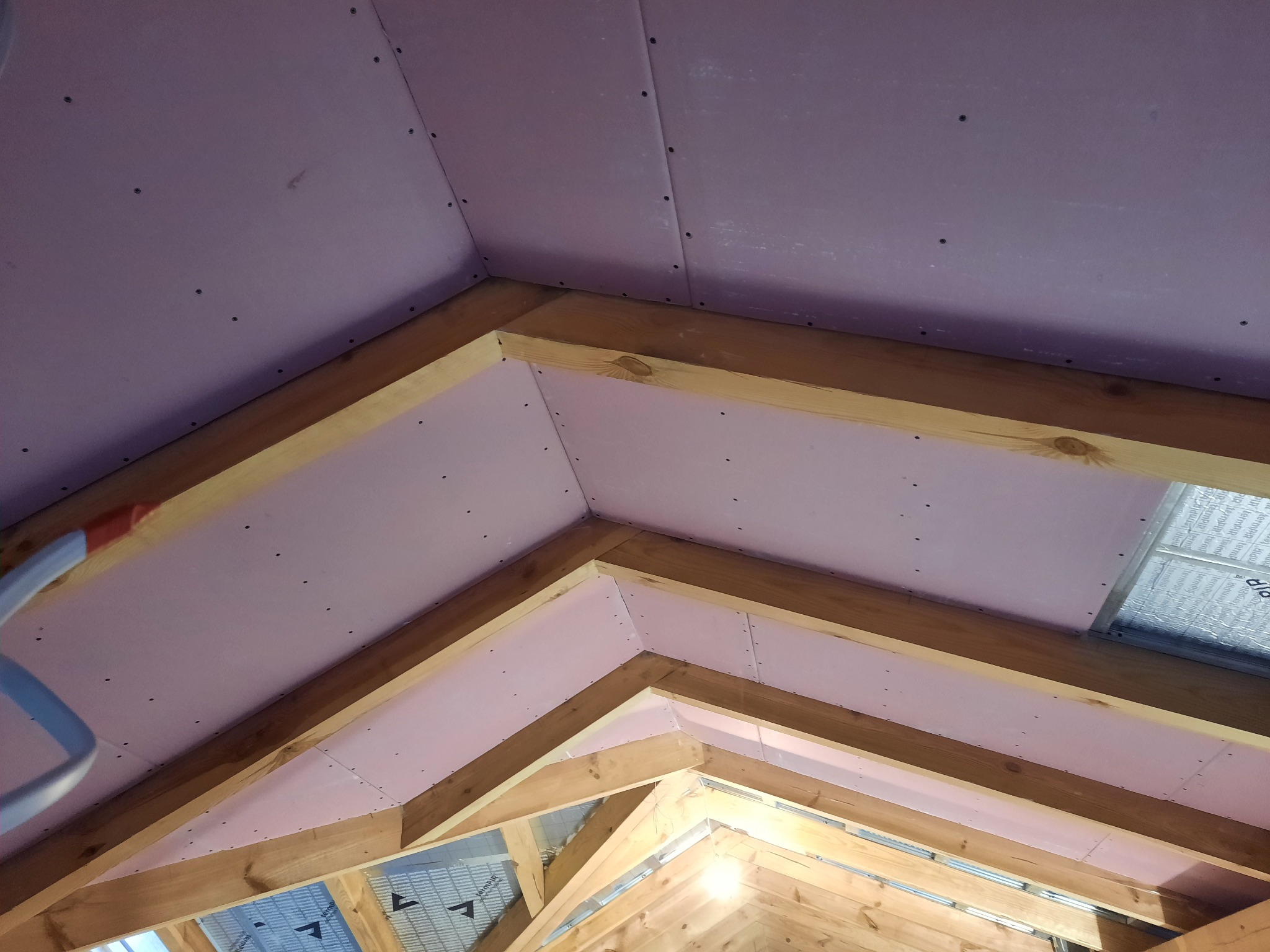 Montaż różowych płyt na drewnianej konstrukcji poddasza, widoczne wkręty i izolacja z folii aluminiowej.
