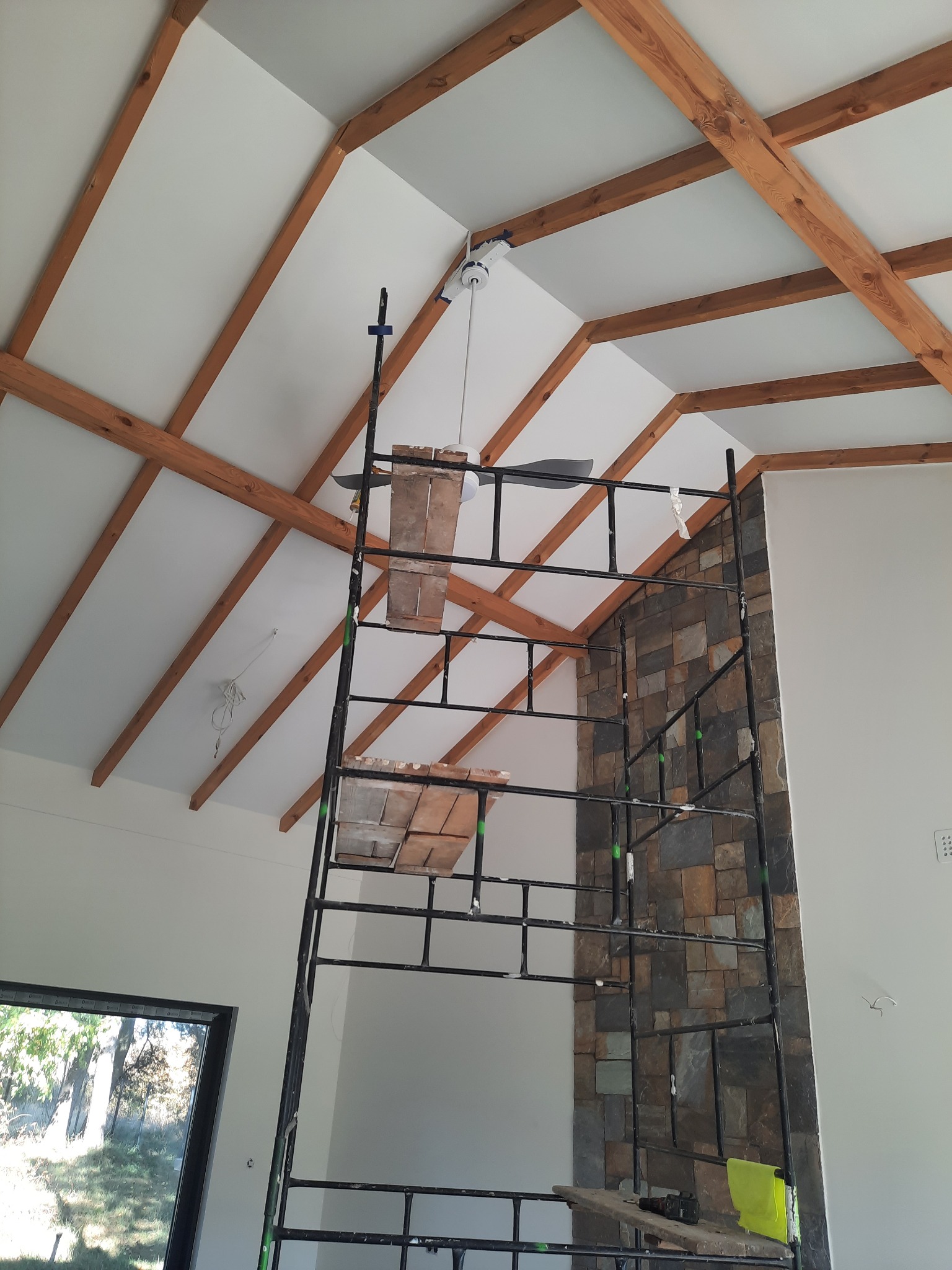 Wnętrze w trakcie prac wykończeniowych: widoczne drewniane belki stropowe, fragment ściany wyłożonej kamieniem dekoracyjnym oraz rusztowanie z drewnianymi podestami.