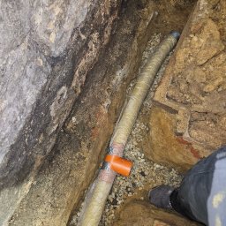 Jeziorski-instalacje Wod-Kan-Gaz-CO roboty ziemne - Rura kanalizacyjna owinięta izolacją, ułożona w wykopie z kamienistym podsypem, widok z góry na fragment nogi w roboczym obuwiu.