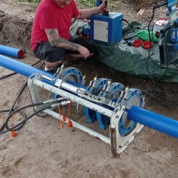 Jeziorski-instalacje Wod-Kan-Gaz-CO roboty ziemne - Pracownik w czerwonej koszulce łączy rury z tworzywa za pomocą zgrzewarki na budowie, widoczna ziemia i fragment zieleni w tle.