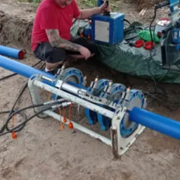 Pracownik w czerwonej koszulce łączy rury z tworzywa za pomocą zgrzewarki na budowie, widoczna ziemia i fragment zieleni w tle.