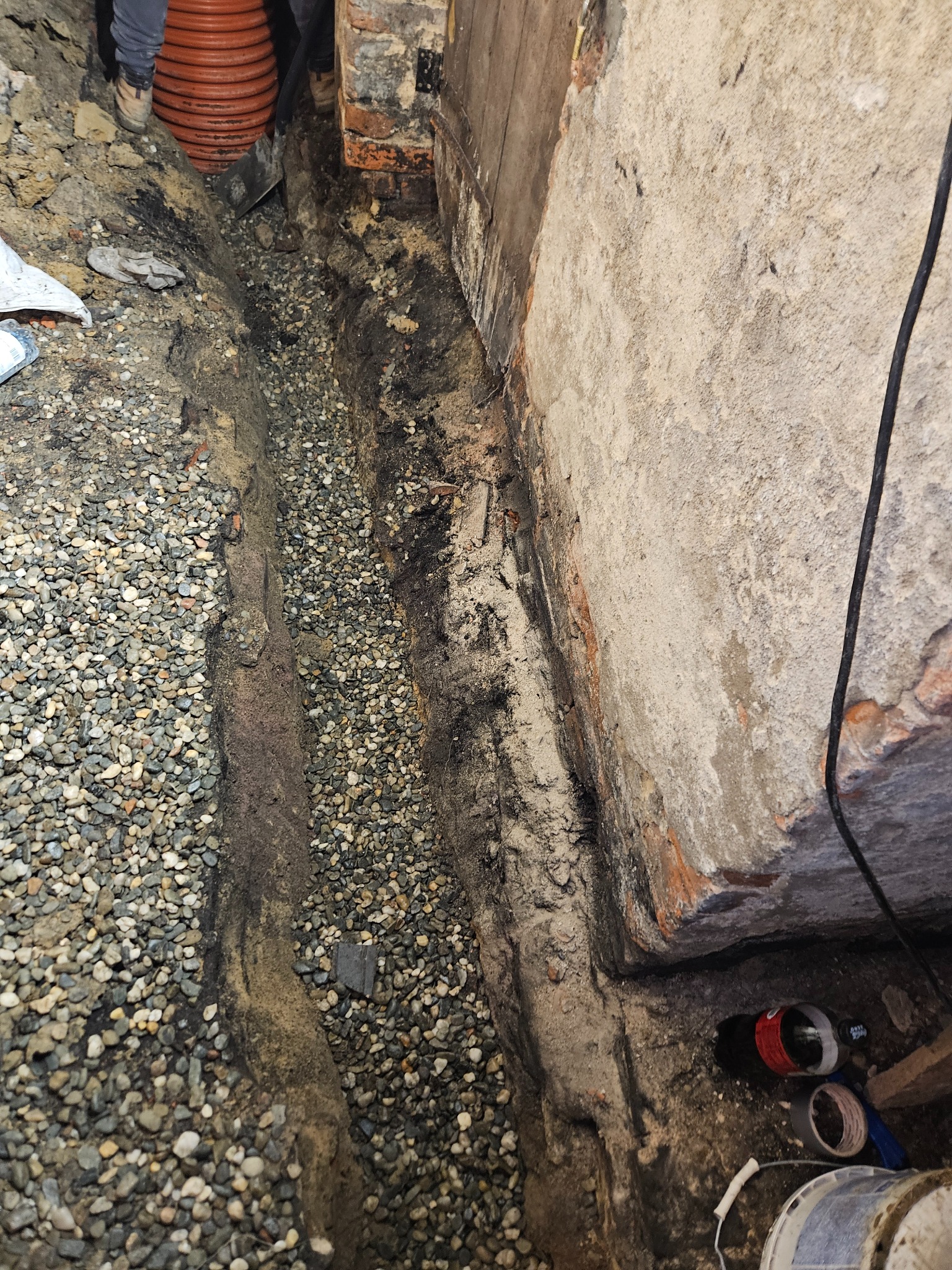 Wykop pod fundamentem z ułożonym drenażem żwirowym, widoczna rura drenarska i fragment ściany z ceglanymi elementami.