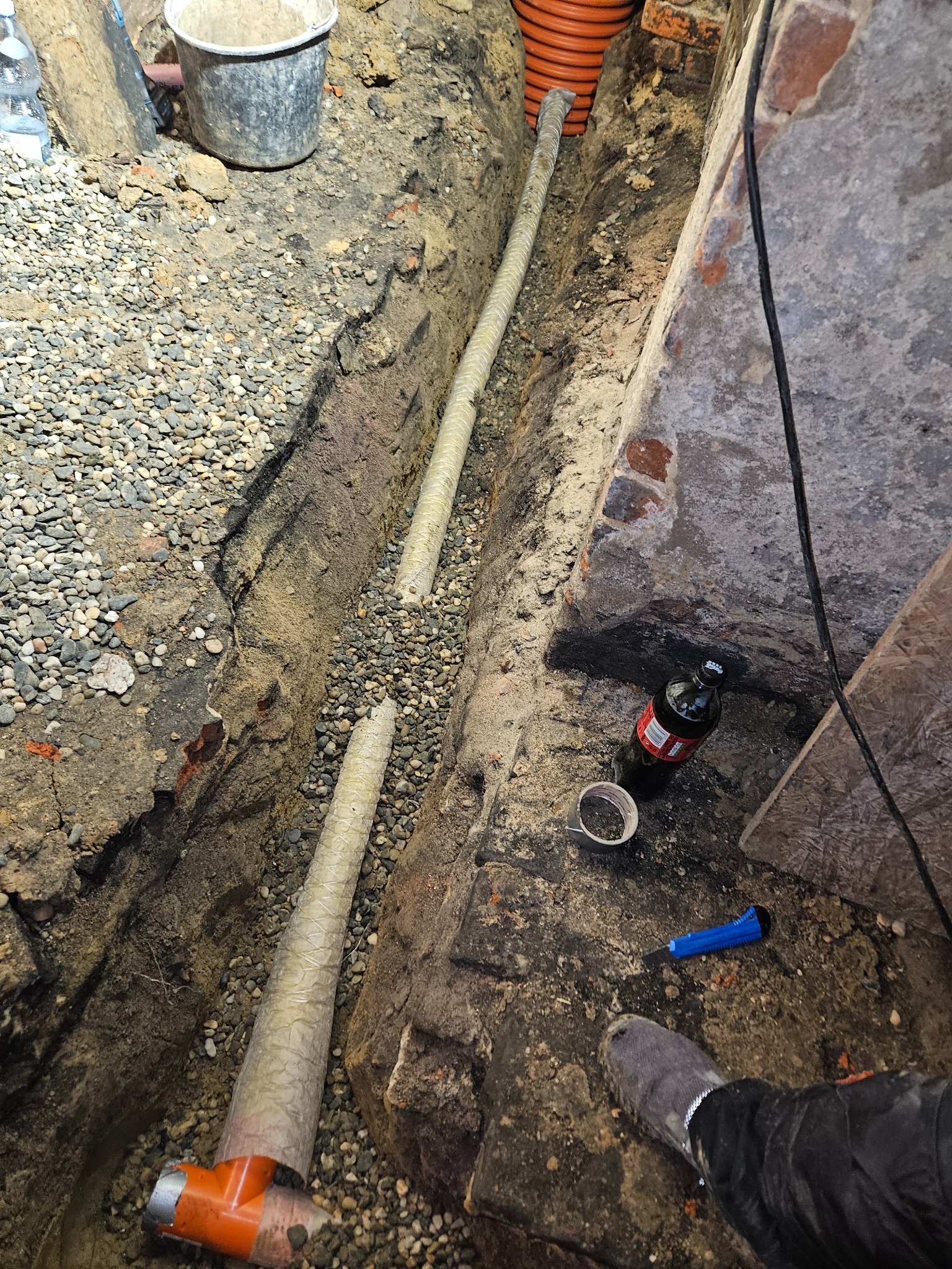 Wykop z dwoma rurami kanalizacyjnymi w osłonie, zasypany częściowo żwirem, widoczna butelka napoju i nóż introligatorski.