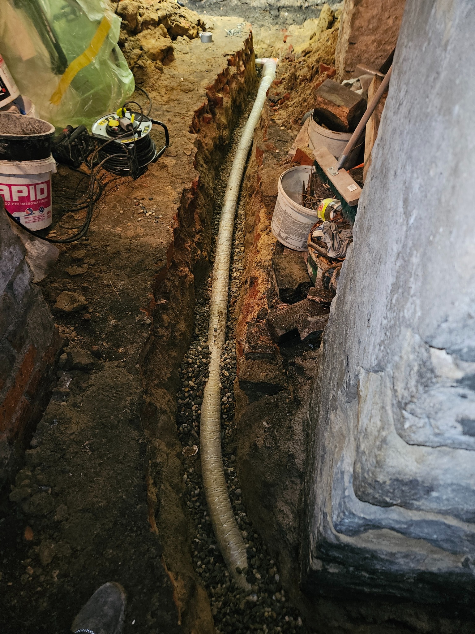 Wykop w ziemi z ułożoną, karbowaną rurą drenażową na warstwie żwiru, widoczne ściany wykopu z ziemi i cegieł oraz fragment buta pracownika w dolnym rogu.