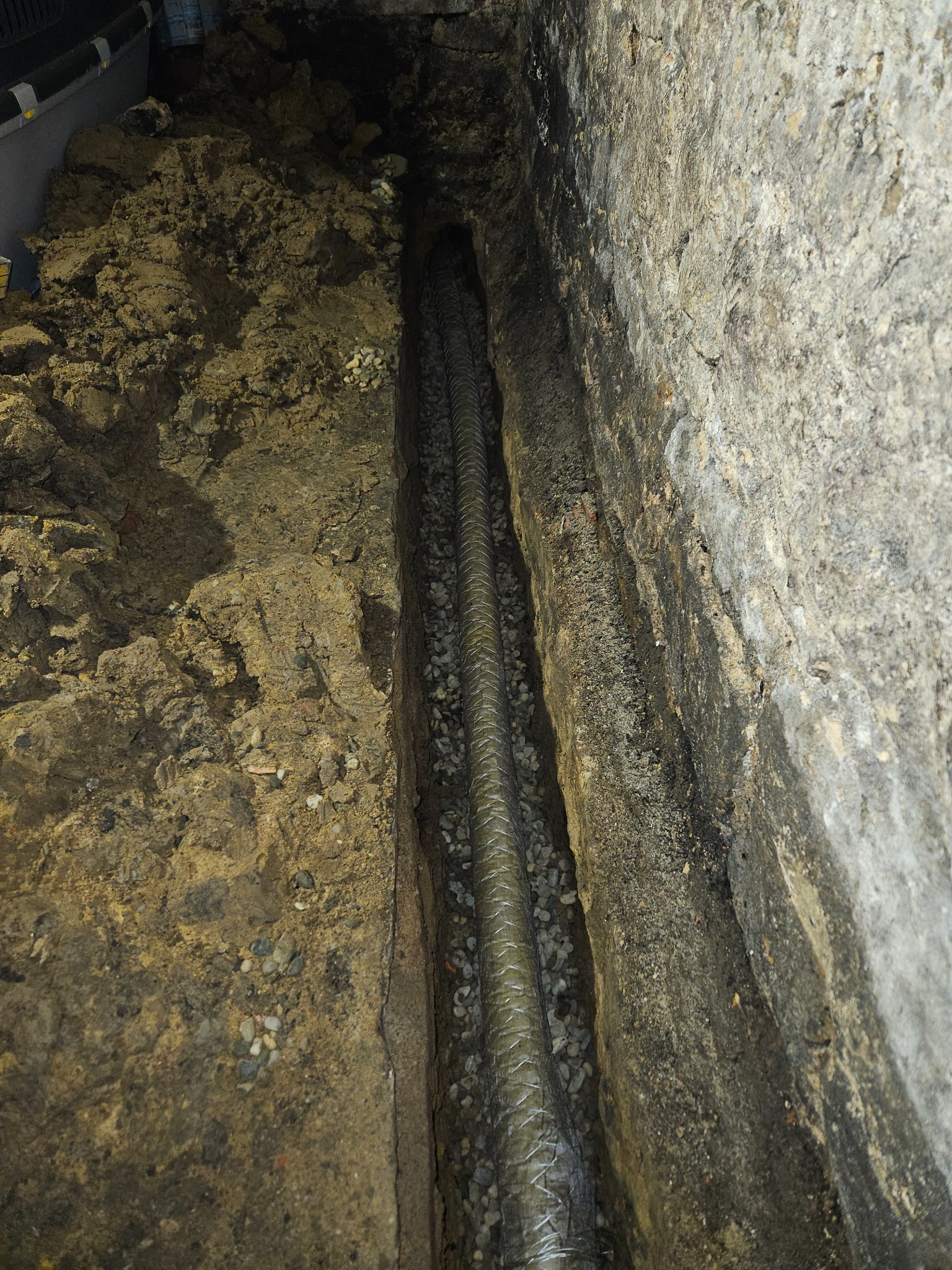 Rów w ziemi z ułożoną rurą w oplocie, zasypaną drobnym żwirem, widoczne ściany wykopu z ziemi i kamienia.