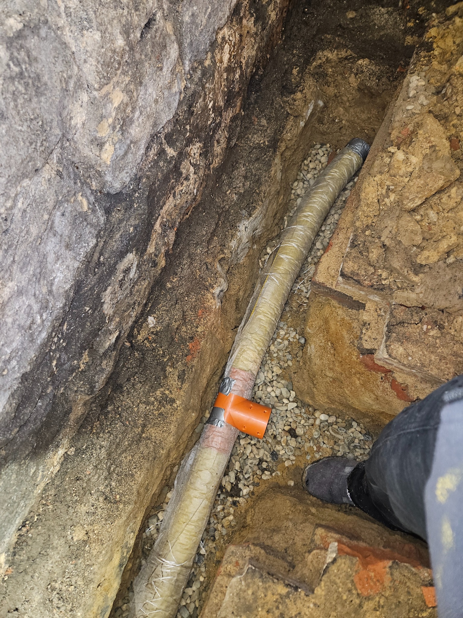 Rura kanalizacyjna owinięta izolacją, ułożona w wykopie z kamienistym podsypem, widok z góry na fragment nogi w roboczym obuwiu.