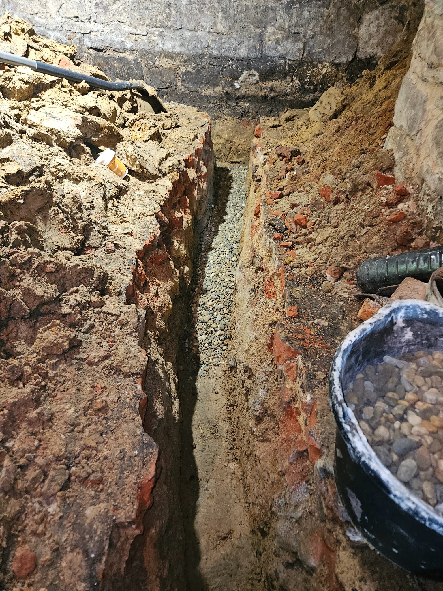 Wykop w ziemi z odsłoniętymi cegłami i rurą, wypełniony drobnym żwirem, z widocznym wiadrem z żwirem w rogu, ukazujący etap prac ziemnych związanych z instalacjami.