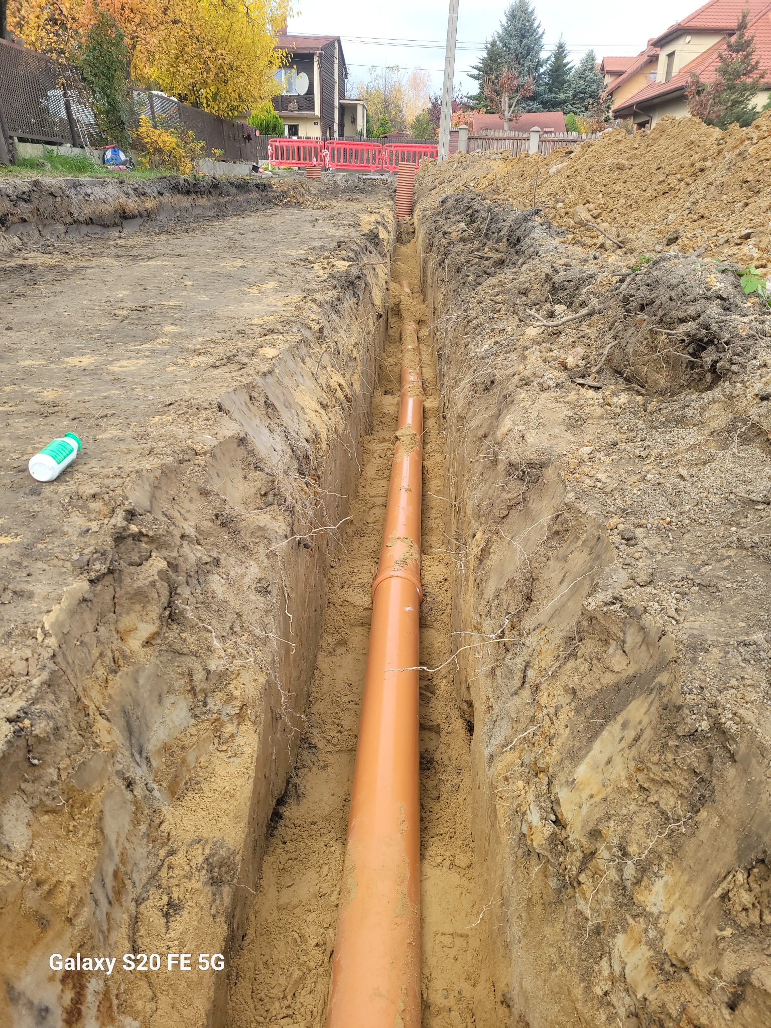 Pomarańczowa rura kanalizacyjna ułożona w wykopie ziemnym, widoczne ślady po wykopie i butelka z preparatem na skraju wykopu.