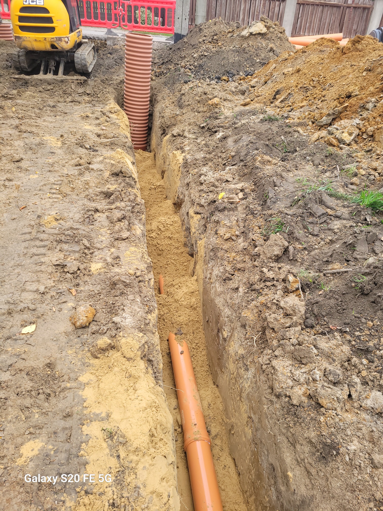 Wykop pod rury kanalizacyjne z widocznym minikoparką JCB w tle, rury z tworzywa sztucznego w kolorze pomarańczowym ułożone w wykopie, ziemia usypana po bokach wykopu.