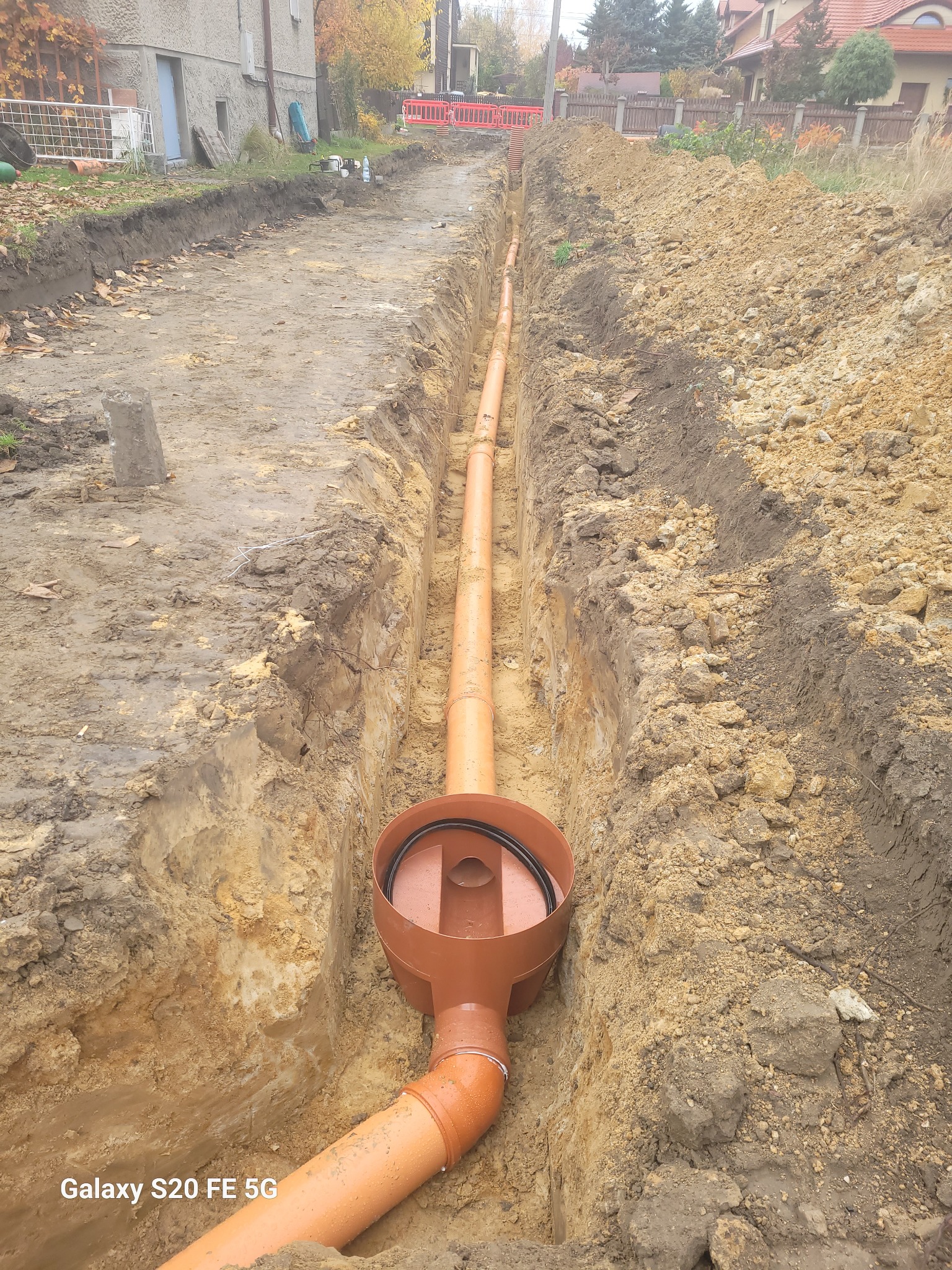 Wykonany wykop z ułożonym pomarańczowym rurociągiem z tworzywa sztucznego i studzienką rewizyjną, widoczne ślady świeżych prac ziemnych.