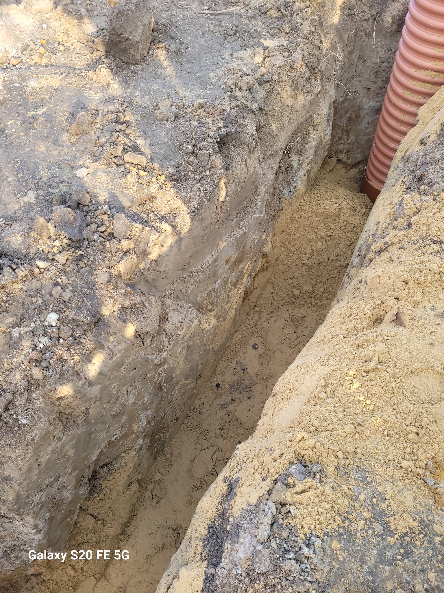 Wykop w ziemi z widoczną rurą kanalizacyjną z tworzywa sztucznego o karbowanej powierzchni, ułożoną wzdłuż jednej ze ścian wykopu.