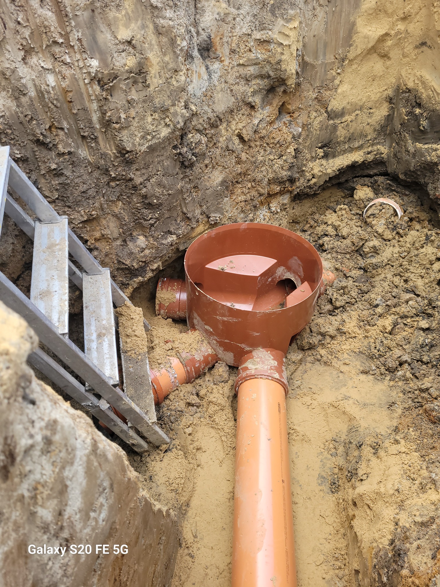 Brązowe rury kanalizacyjne z trójnikiem w wykopie ziemnym, drabina aluminiowa oparta o krawędź wykopu, widoczne ślady wody w rurze.