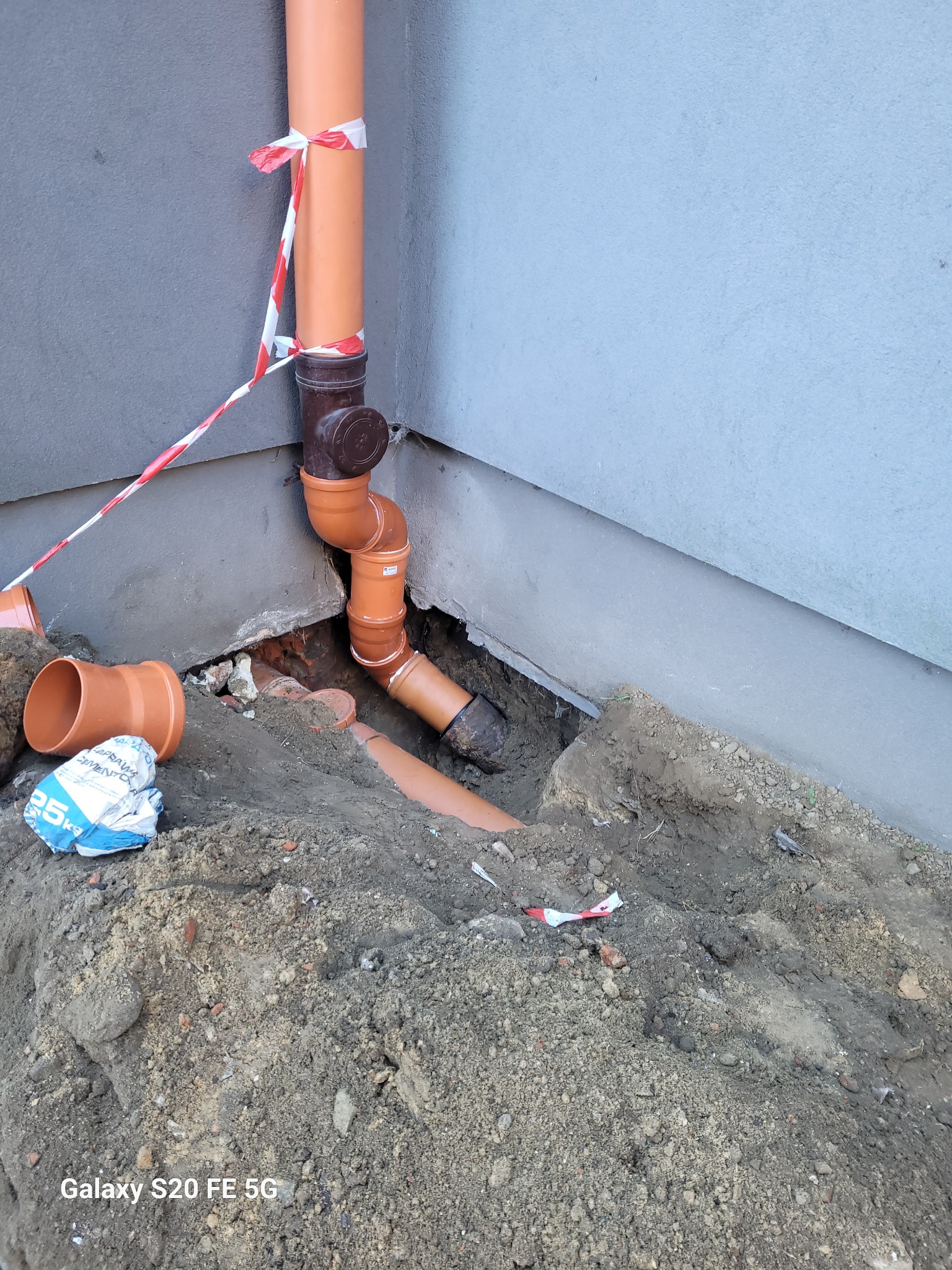 Instalacja rur kanalizacyjnych z pomarańczowego PCV, częściowo zakopanych w ziemi przy fundamencie budynku, zabezpieczonych taśmą ostrzegawczą.