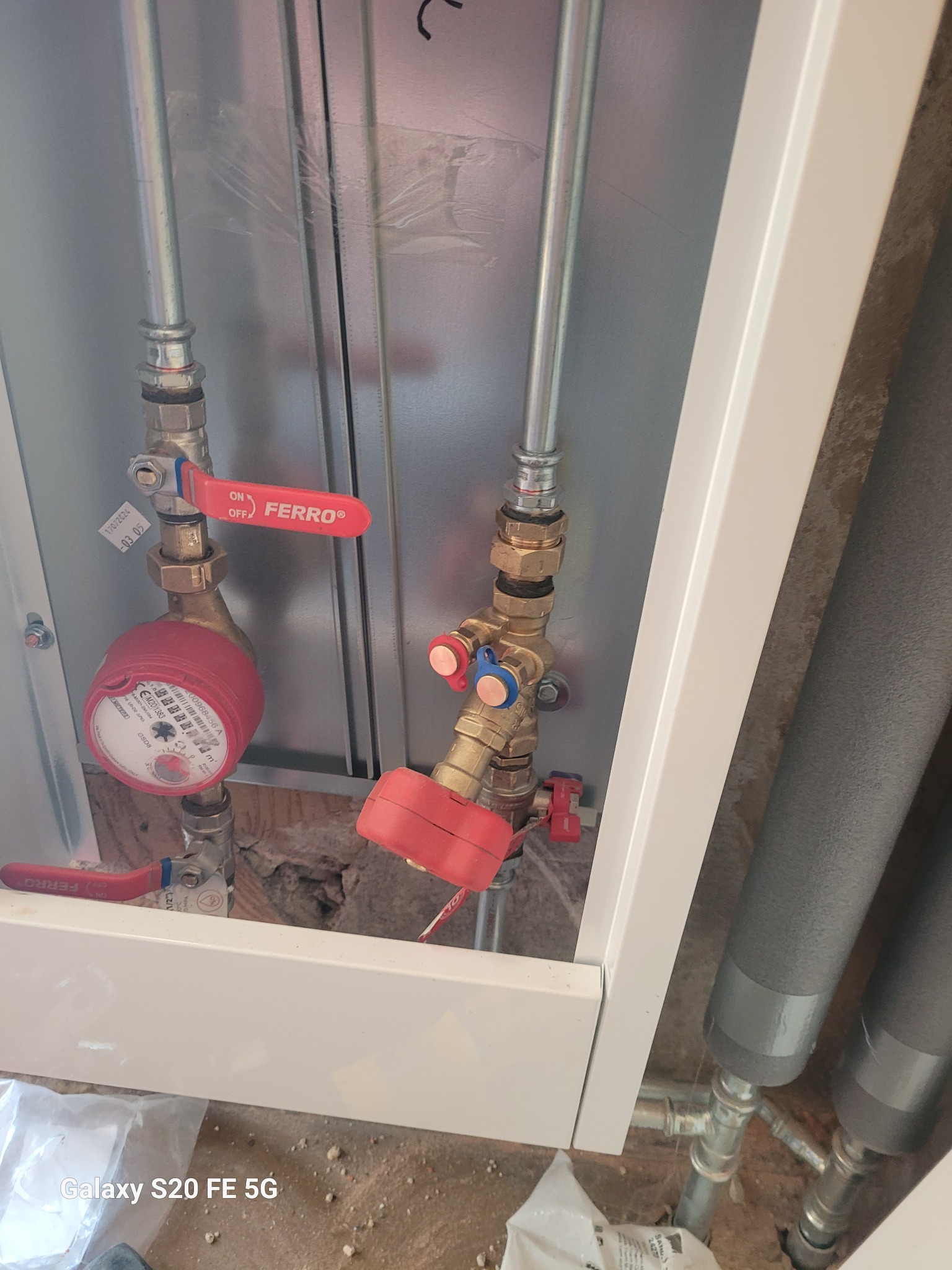 Wewnętrzna instalacja wodna z licznikiem i zaworami kulowymi marki Ferro, widoczne rury stalowe i izolowane rury, montaż w szafce z białymi elementami wykończeniowymi.