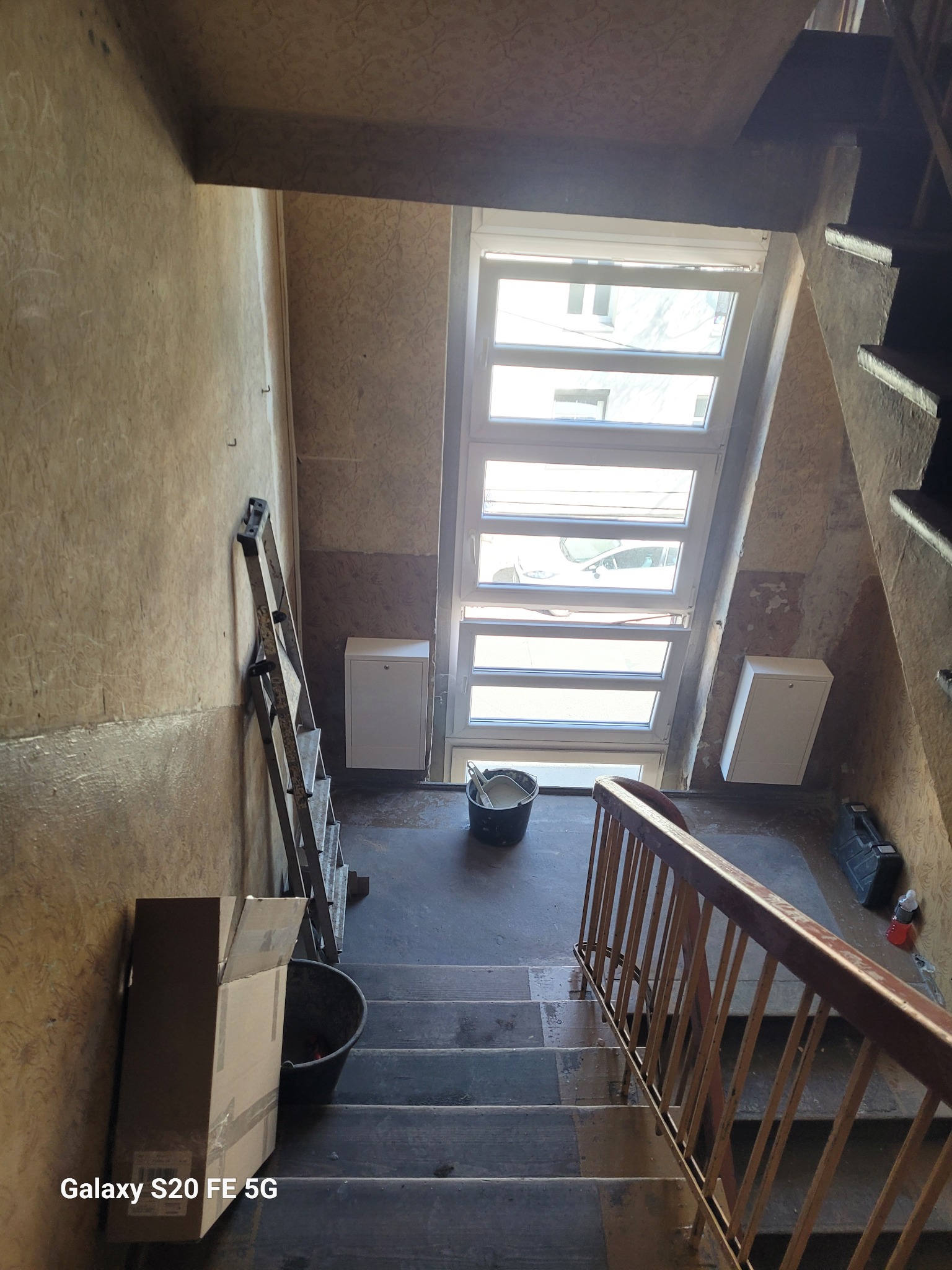 Klatka schodowa w trakcie remontu: drabina oparta o ścianę, wiadro z zaprawą, skrzynka narzędziowa i okno doświetlające. Widoczne ślady po demontażu elementów instalacji na ścianie.