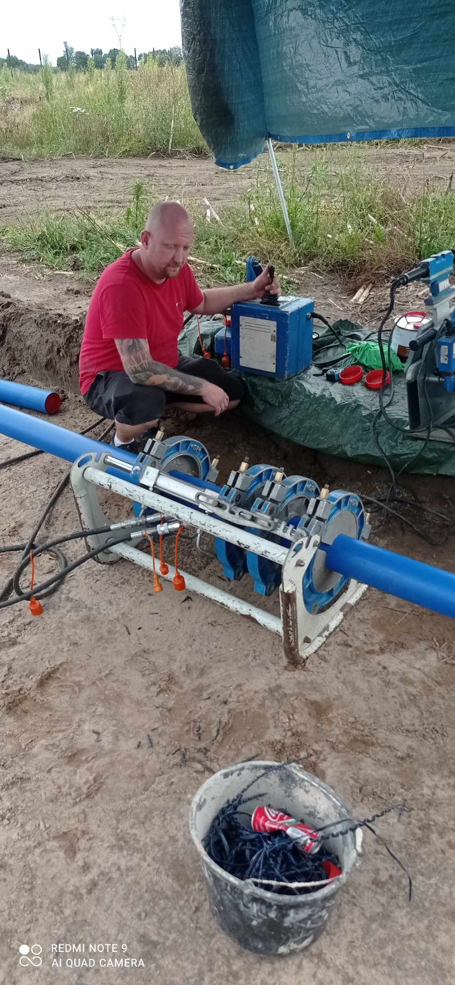 Pracownik w czerwonej koszulce łączy rury z tworzywa za pomocą zgrzewarki na budowie, widoczna ziemia i fragment zieleni w tle.