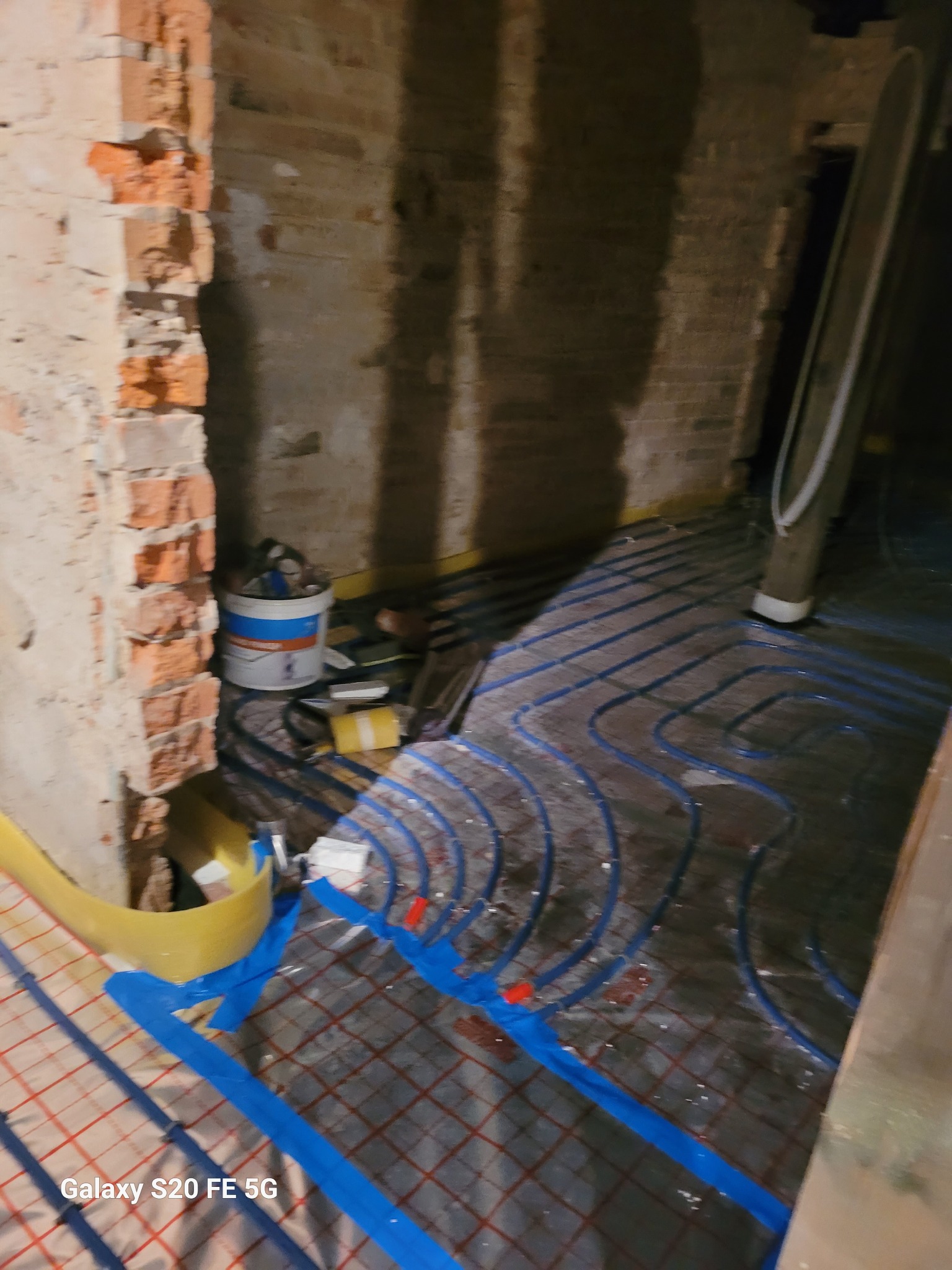 Instalacja ogrzewania podłogowego w trakcie prac, widoczne rury w kształcie meandrów, mocowane taśmą do podłoża z siatką, fragment ściany z cegły, wiadro z klejem i narzędzia w tle.