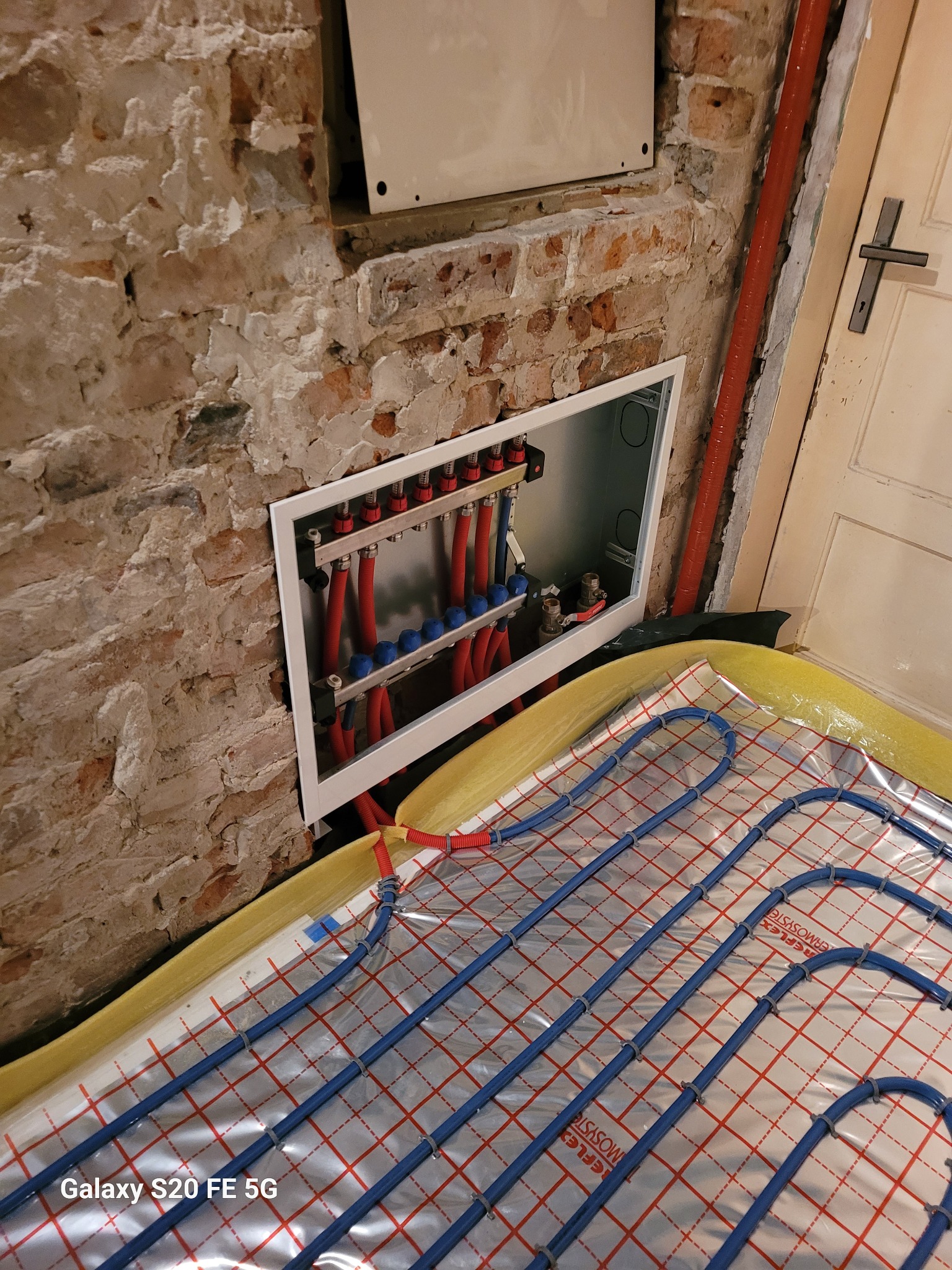 Rozdzielacz ogrzewania podłogowego zamontowany na surowej ścianie z cegły, widoczne niebieskie rurki instalacji ułożone na folii izolacyjnej z żółtymi krawędziami.