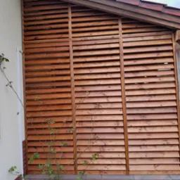 Drewniana konstrukcja z poziomych listew, tworząca osłonę na tarasie, z widocznym fragmentem dachu i pnącą rośliną na pierwszym planie.