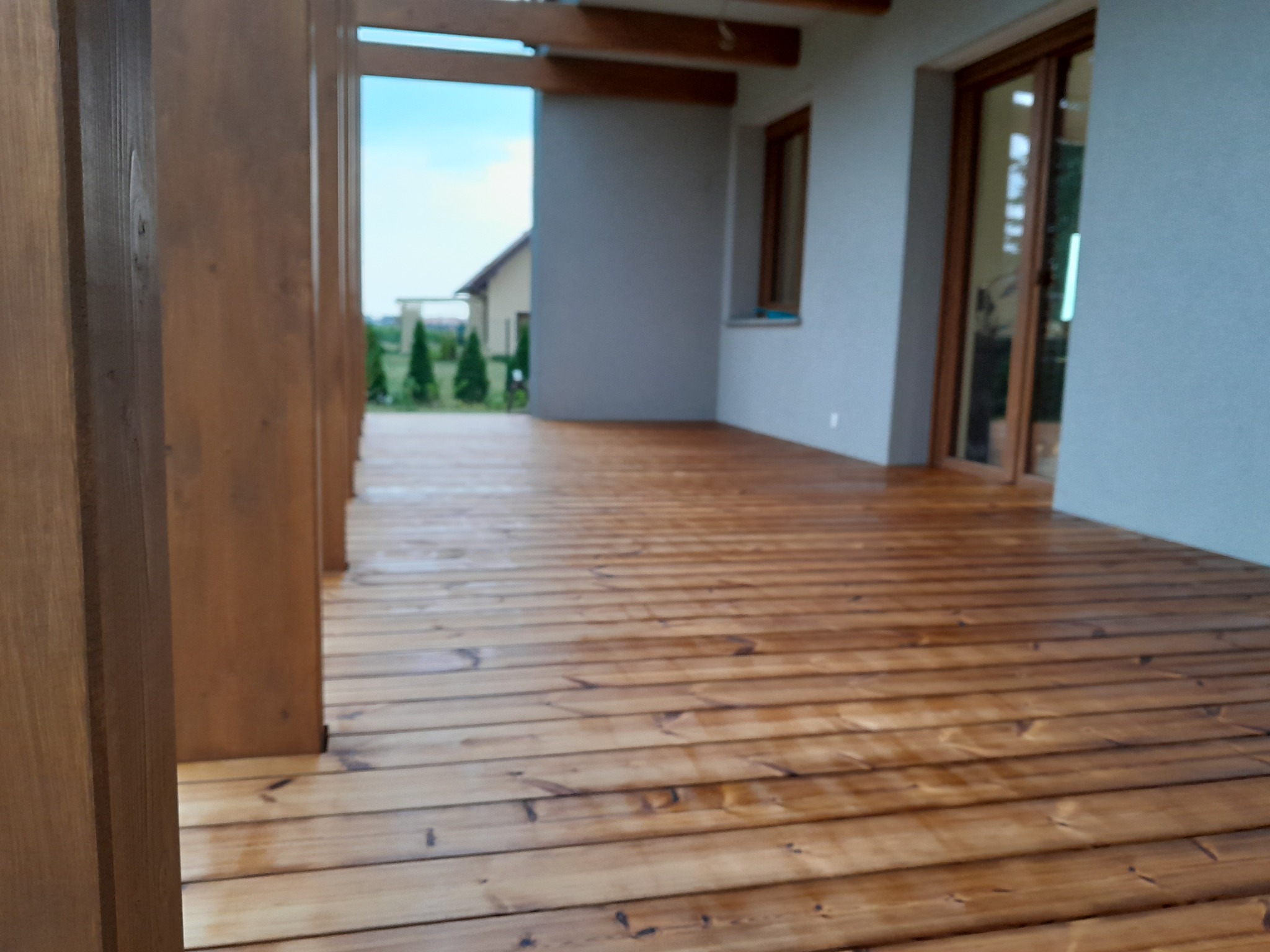 Zadaszony taras z impregnowanej deski, widok na ogród przez drewnianą konstrukcję, szara elewacja budynku w tle.