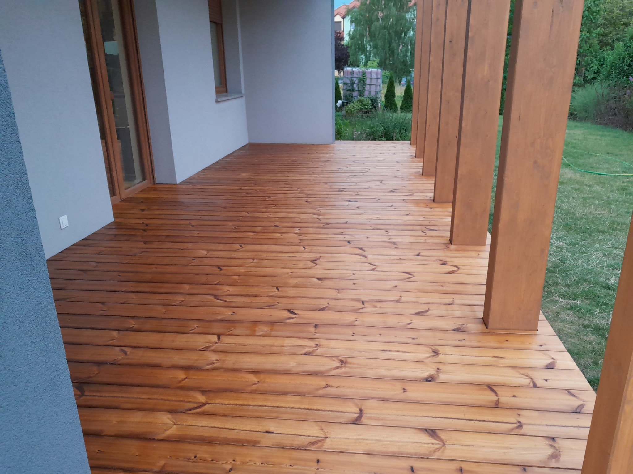 Nowoczesny taras z desek w kolorze miodowego brązu, z widocznymi słupami konstrukcyjnymi i fragmentem elewacji budynku w tle.