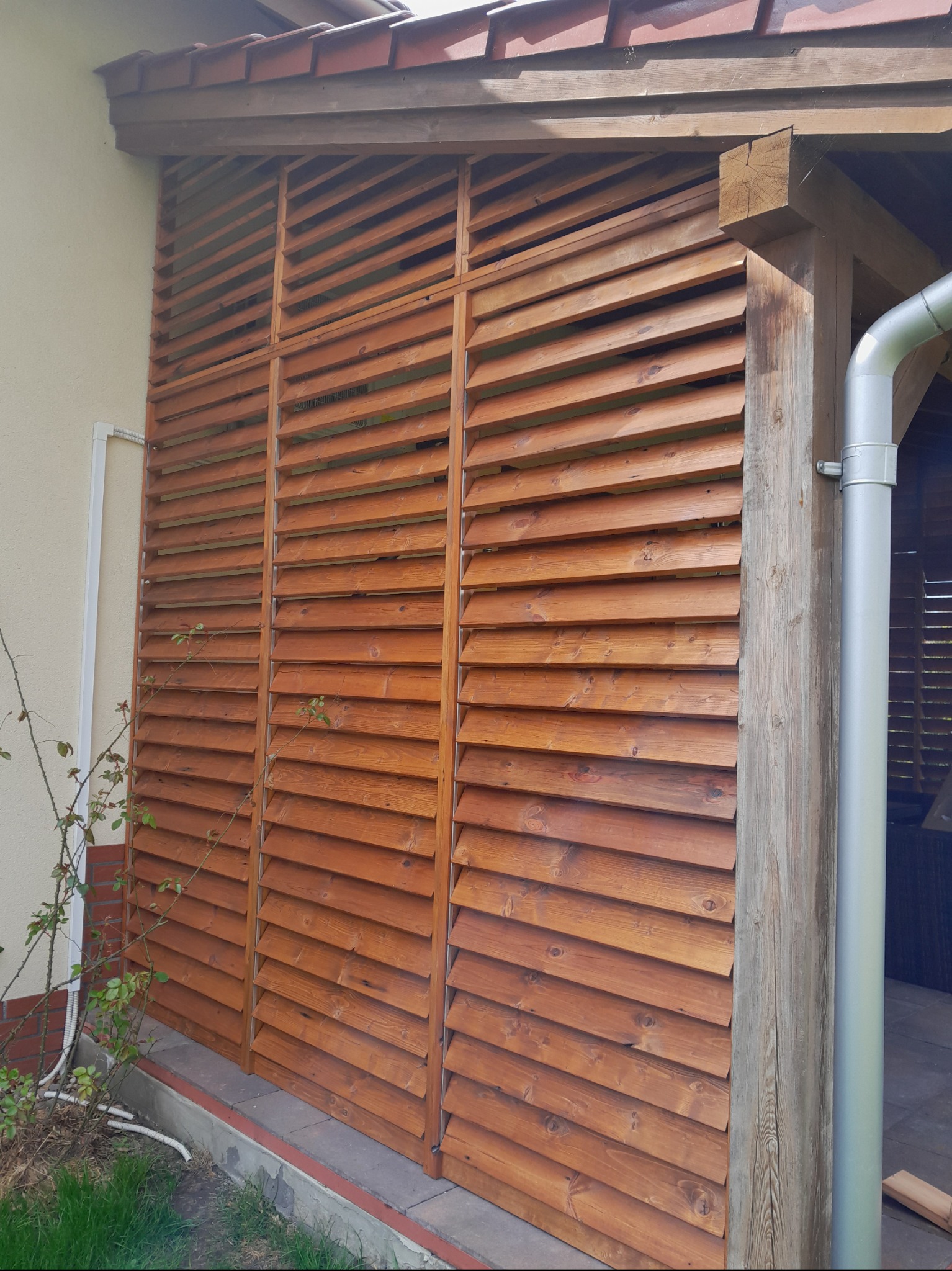 Zewnętrzna konstrukcja z drewnianych żaluzji, prawdopodobnie fragment pergoli lub altany, z widocznym słupem nośnym i rynną spustową, przylegająca do ściany budynku z fragmentem instalacji...