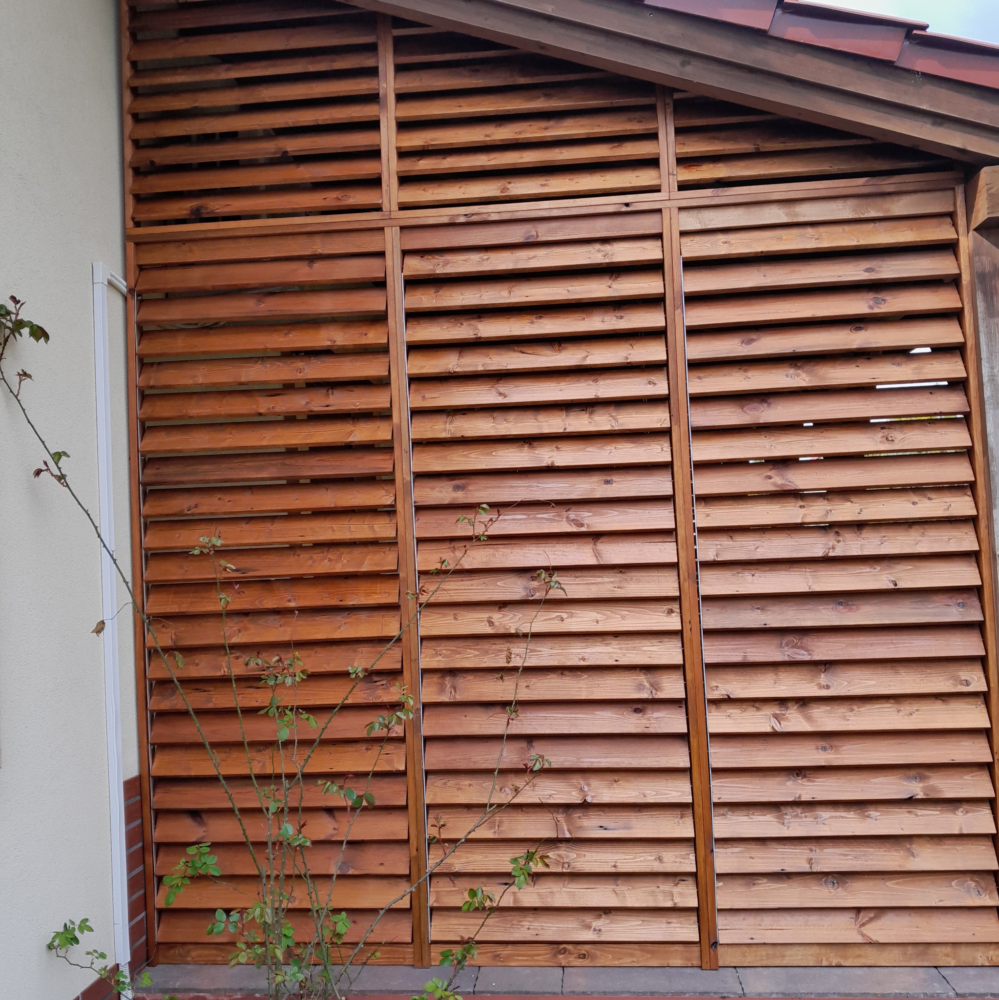 Drewniana konstrukcja z poziomych listew, tworząca osłonę na tarasie, z widocznym fragmentem dachu i pnącą rośliną na pierwszym planie.