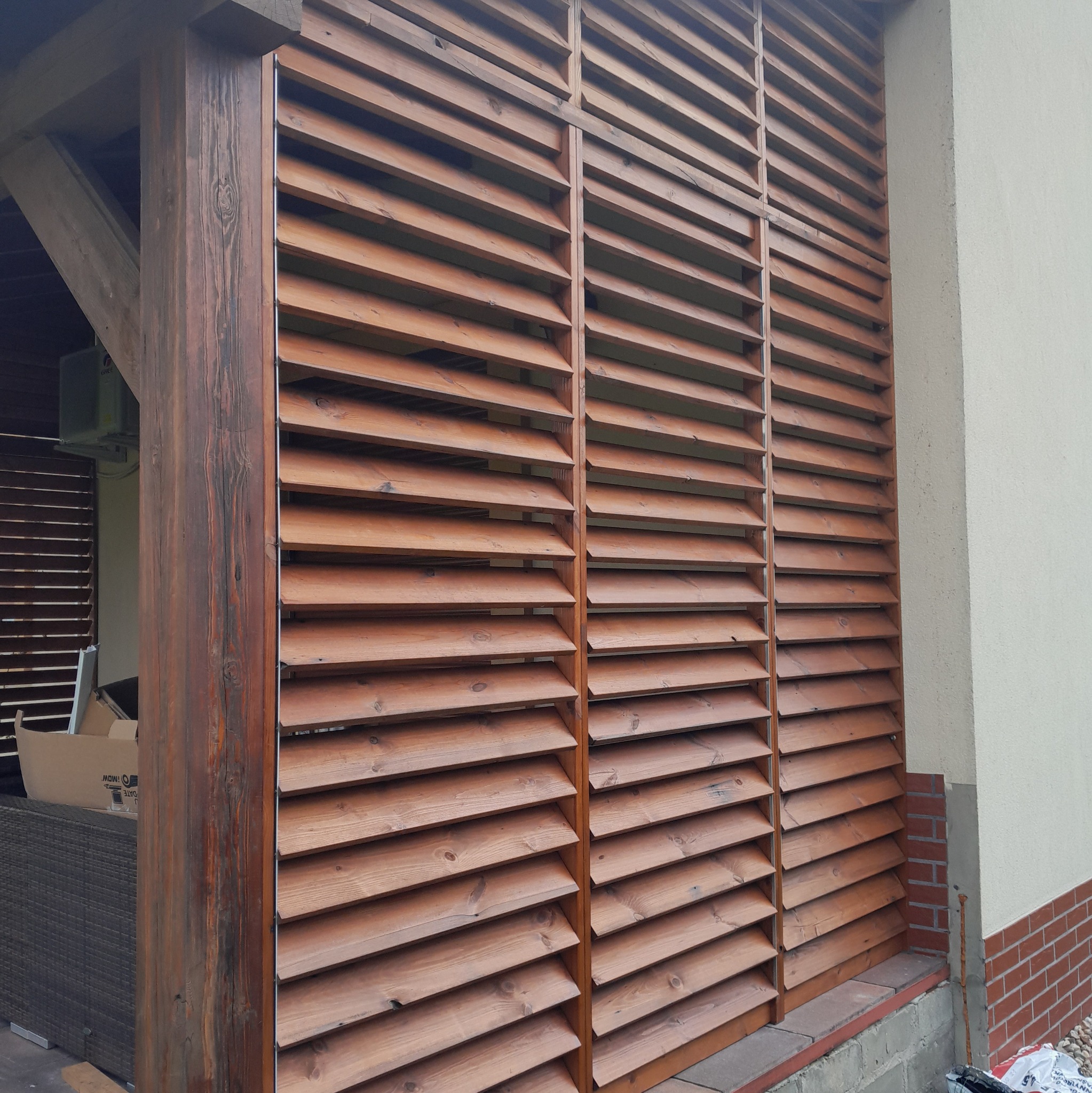 Drewniana pergola z lamelami, tworząca osłonę przeciwsłoneczną, przylegająca do ściany budynku, z widoczną konstrukcją nośną z ciemnego drewna.