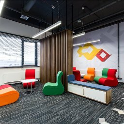 PISAREK MARCIN PRZEDSIĘBIORSTWO USŁUGOWO HANDLOWE PLAST-BUD - Nowoczesne biuro z wykładziną dywanową w odcieniach szarości, kolorowe fotele o nietypowym kształcie, drewniana ścianka działowa, designerski wzór na ścianie i oświetlenie liniowe.