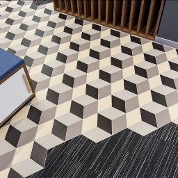 PISAREK MARCIN PRZEDSIĘBIORSTWO USŁUGOWO HANDLOWE PLAST-BUD - Geometryczny wzór podłogi z płytek w odcieniach szarości i beżu, tworzący iluzję trójwymiarowych kostek, przechodzący w szarą wykładzinę dywanową z białymi prążkami, z fragmentem niebieskiej pufy...