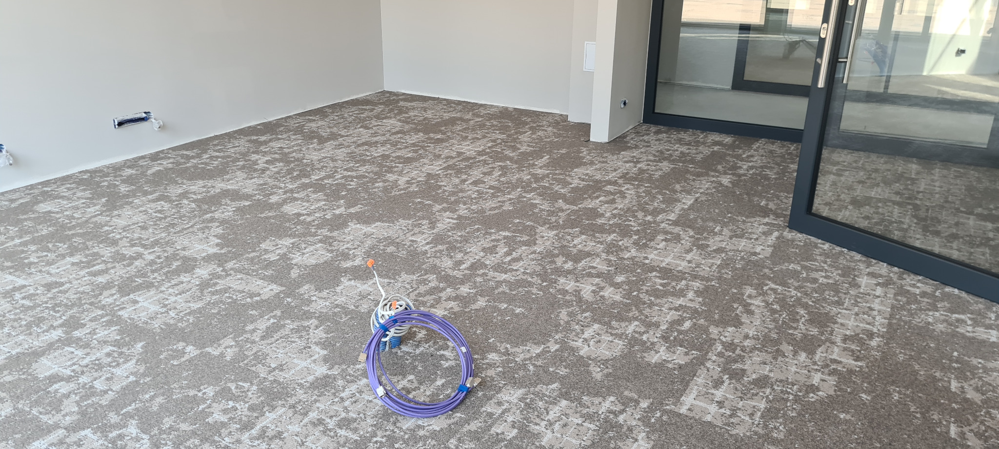 Nowo położona podłoga z wzorzystą, beżowo-szarą powierzchnią w pustym pomieszczeniu z dużym oknem przesuwnym i niepodłączonymi kablami sieciowymi na środku.