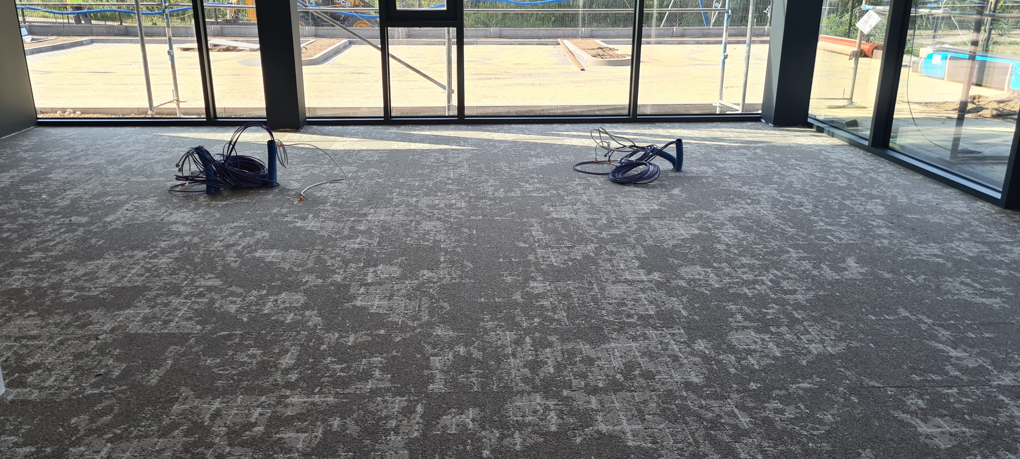 Szara wykładzina dywanowa o abstrakcyjnym wzorze w nowym pomieszczeniu, z widocznymi przewodami elektrycznymi i budową za oknem.