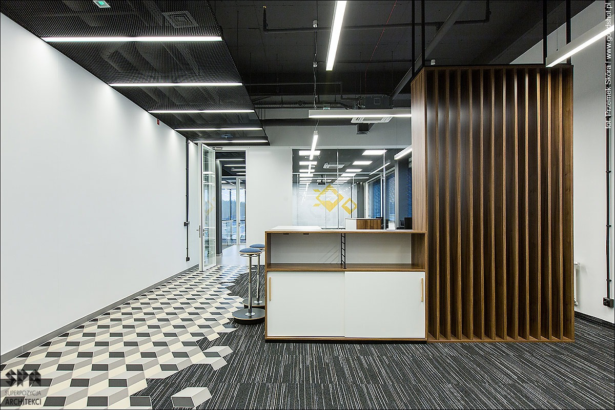 Nowoczesne biuro z wykładziną dywanową w szaro-białe prążki, geometryczna podłoga w strefie wejścia, drewniana ścianka działowa z pionowych listew i białe szafki z przesuwnymi drzwiami.