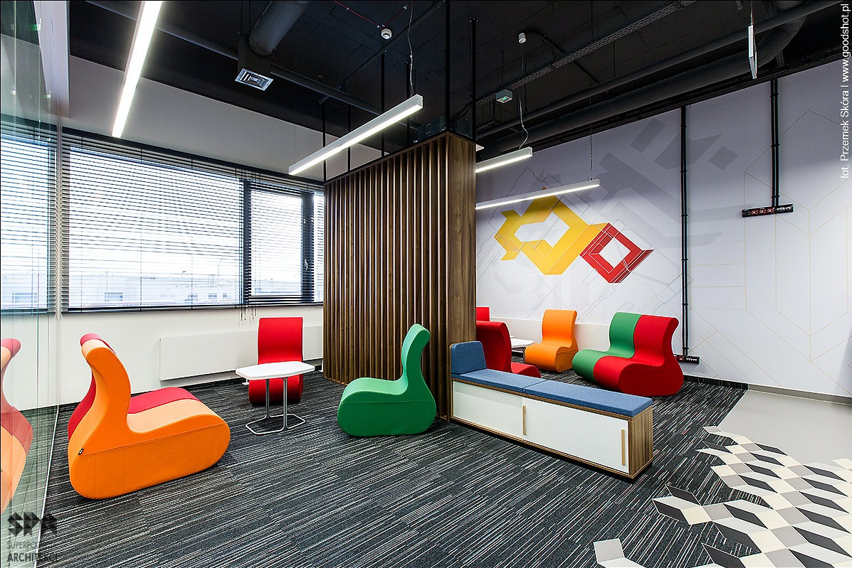 Nowoczesne biuro z wykładziną dywanową w odcieniach szarości, kolorowe fotele o nietypowym kształcie, drewniana ścianka działowa, designerski wzór na ścianie i oświetlenie liniowe.
