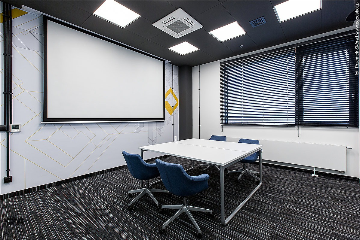 Sala konferencyjna z ciemną wykładziną dywanową w paski, białym stołem i niebieskimi krzesłami na kółkach, z roletami w oknach i ekranem projekcyjnym na ścianie.