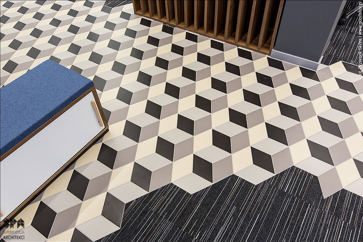 Geometryczny wzór podłogi z płytek w odcieniach szarości i beżu, tworzący iluzję trójwymiarowych kostek, przechodzący w szarą wykładzinę dywanową z białymi prążkami, z fragmentem niebieskiej pufy...